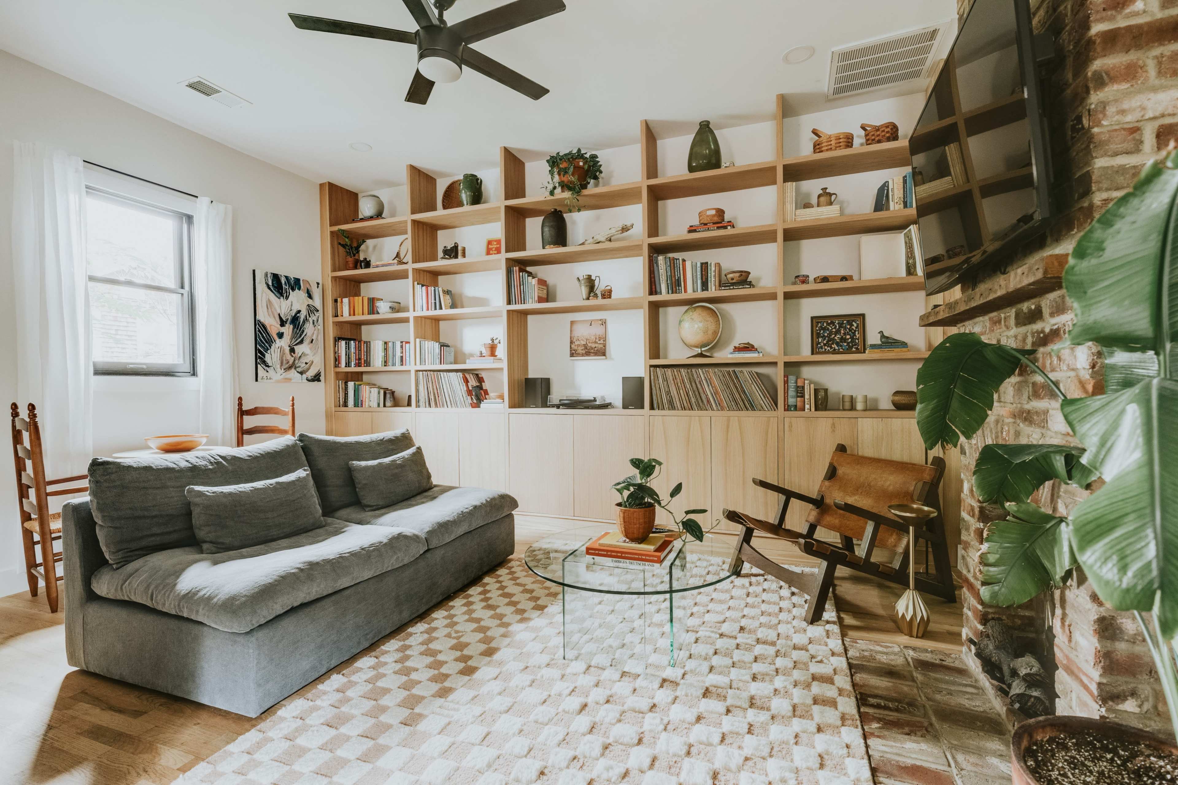A living room with a gray sofa, a glass coffee table, a bookshelf filled with books, and a brick fireplace, all arranged in a well-lit space.