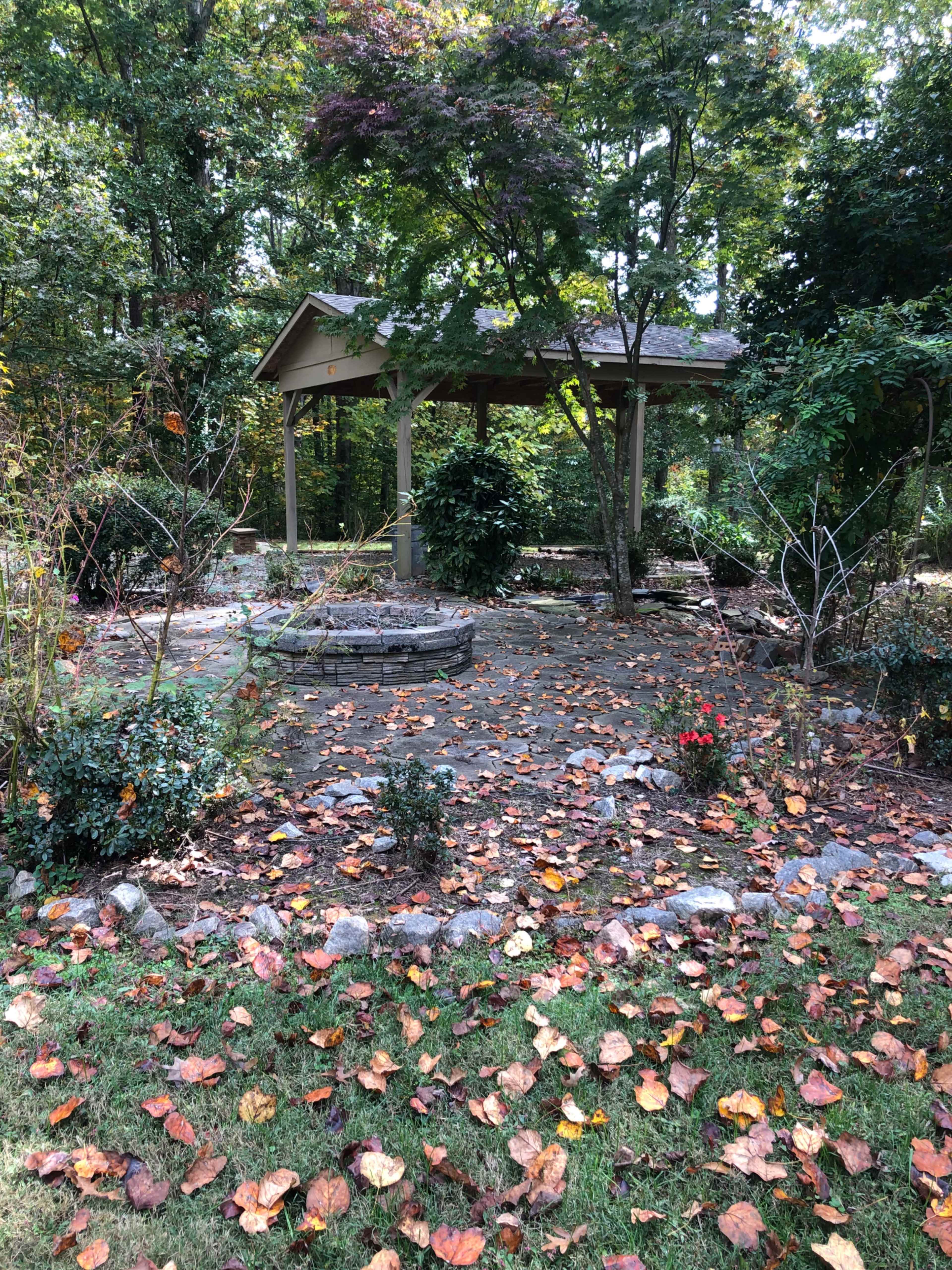 A wooden gazebo is situated in a garden surrounded by trees, with a stone path covered in fallen leaves.