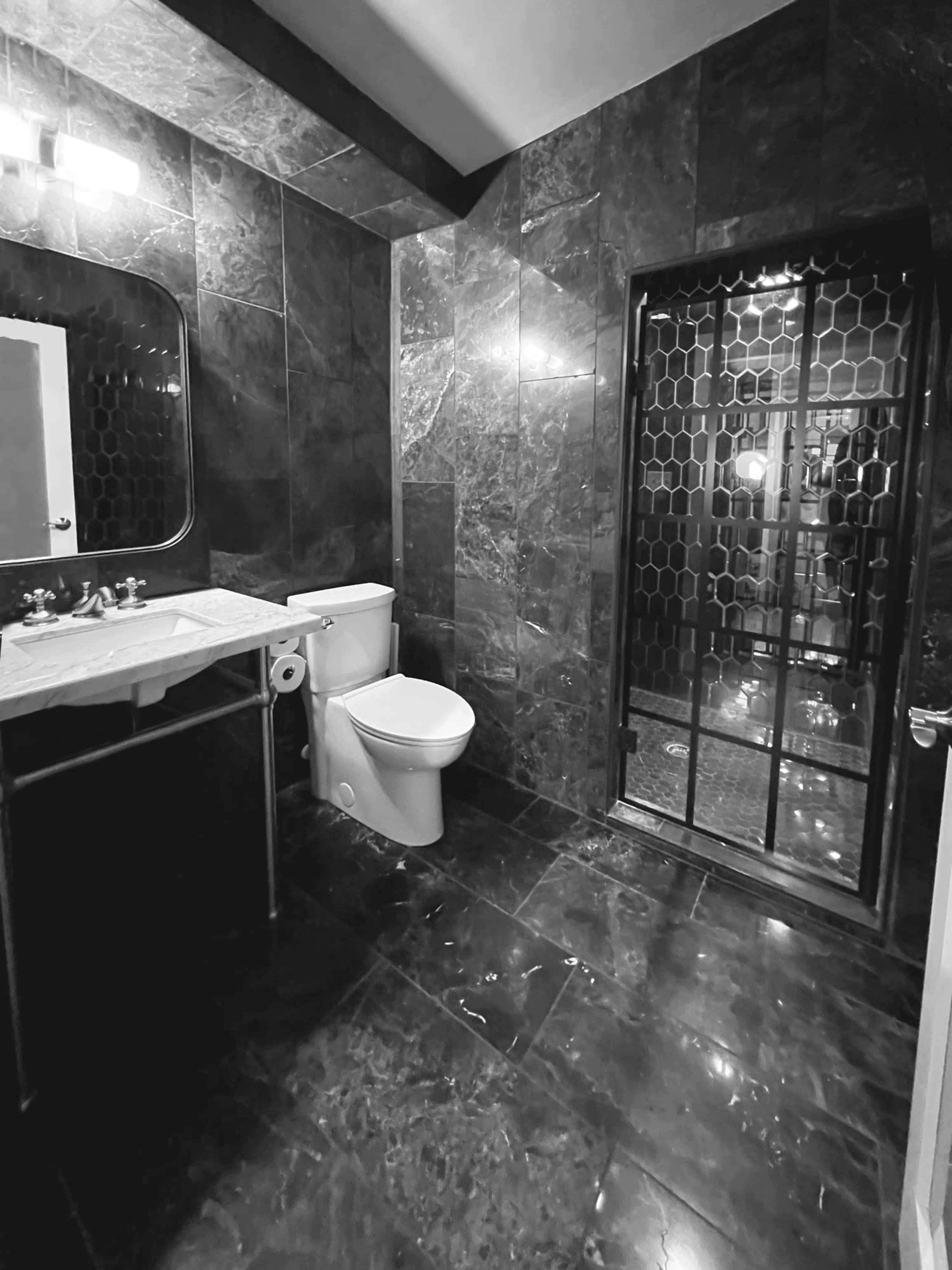 The image shows a bathroom featuring dark marble walls and floor, a toilet, a sink on a metal frame, and a glass shower enclosure.