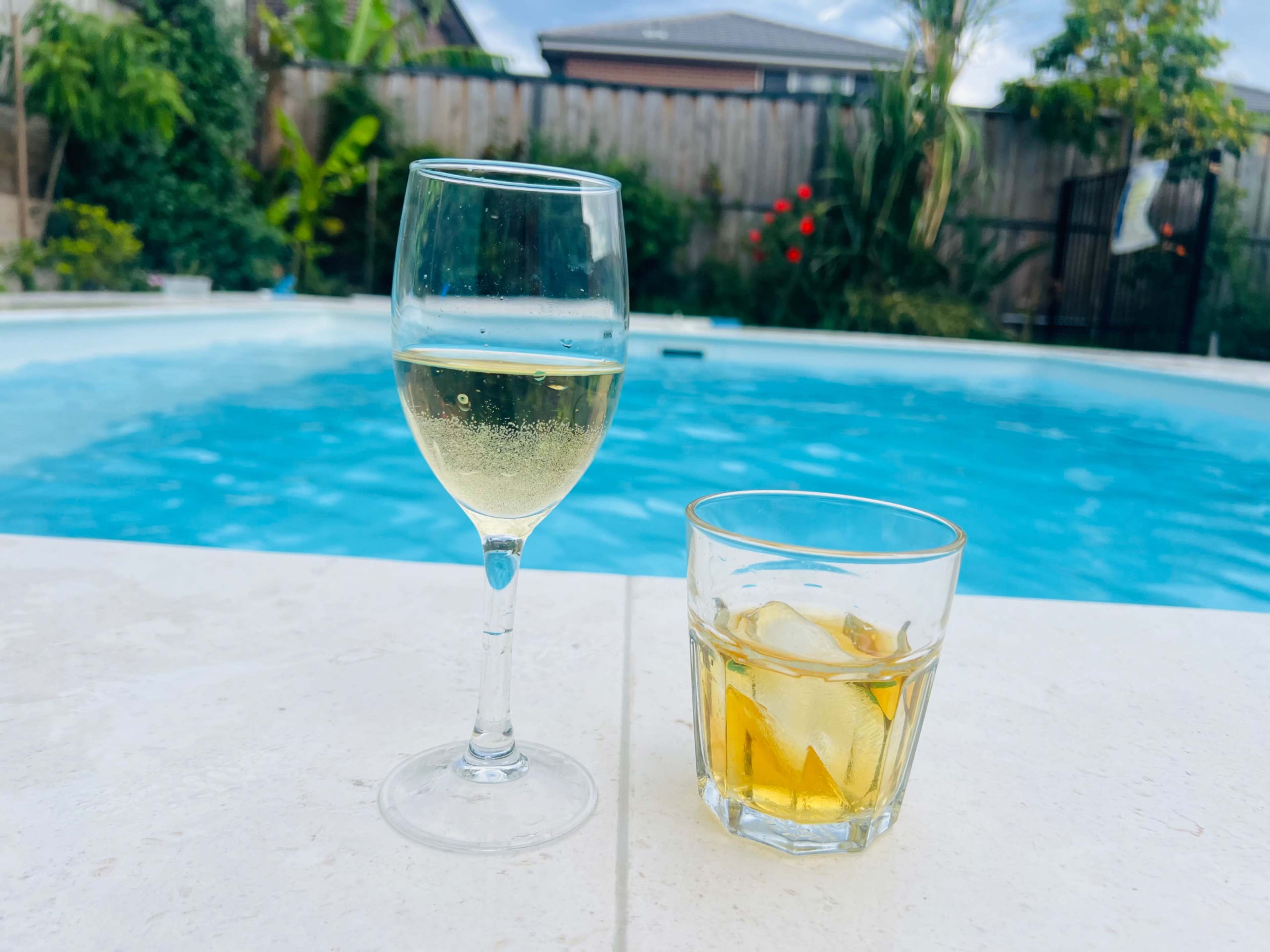 A glass of white wine and a tumbler of whiskey on a marble ledge beside a swimming pool.