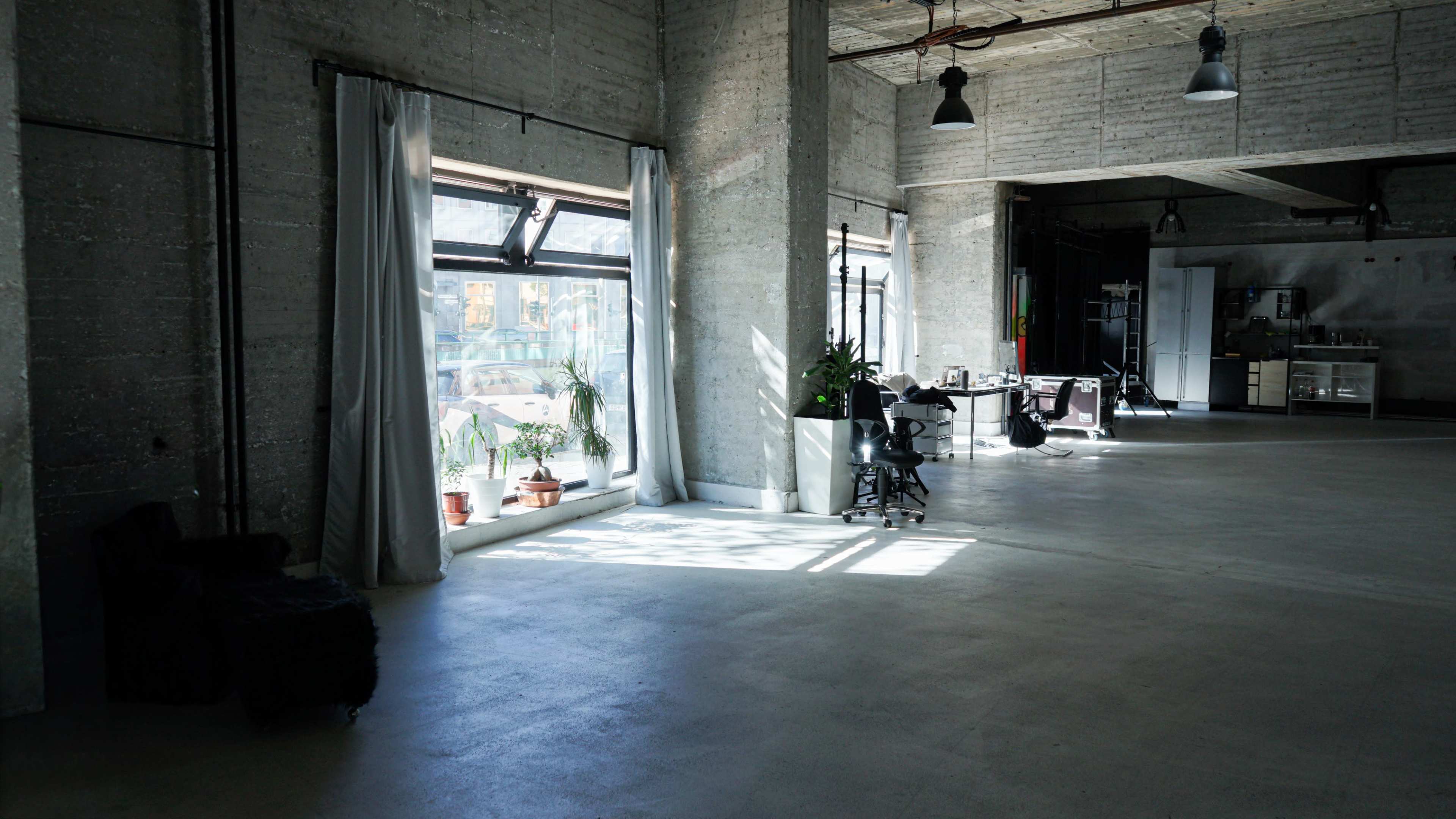 The image shows an industrial-style interior room with large windows, concrete walls, and scattered furniture, including chairs and plants.