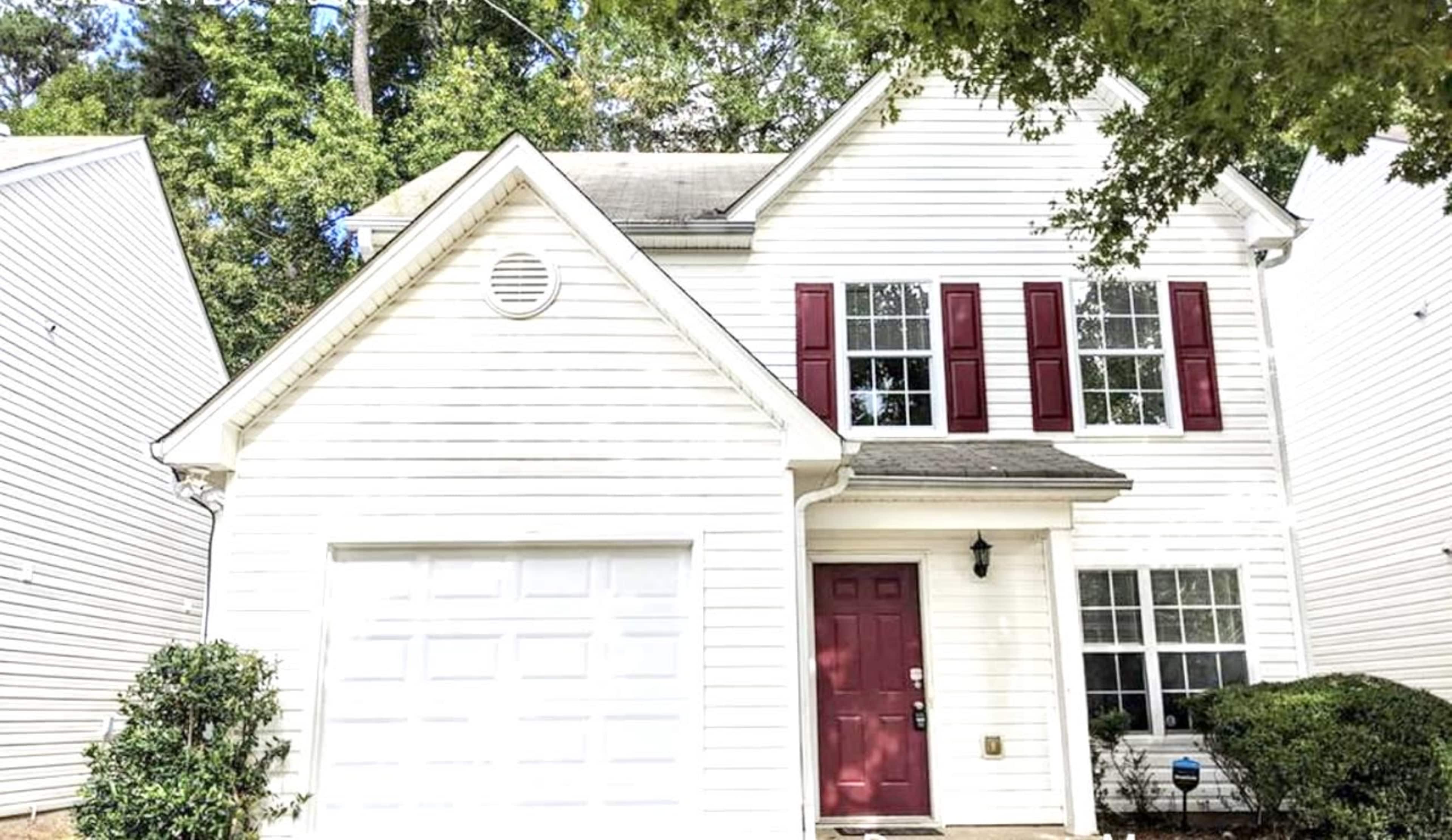 A two-story white house with red shutters and a garage is surrounded by green trees and shrubs.