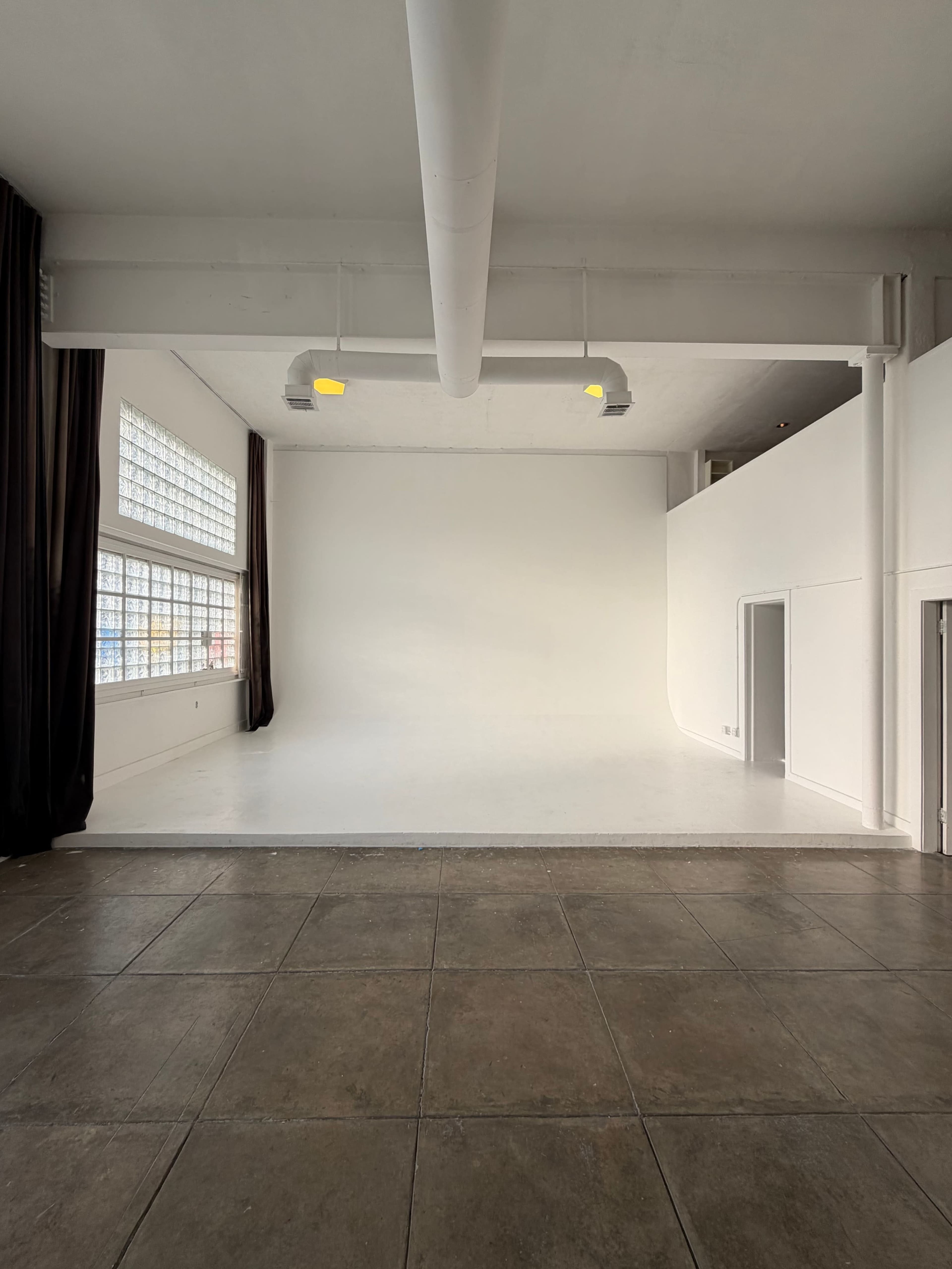 The image shows a spacious, empty room with white walls, a smooth white floor, large windows, and a tiled section on the floor.
