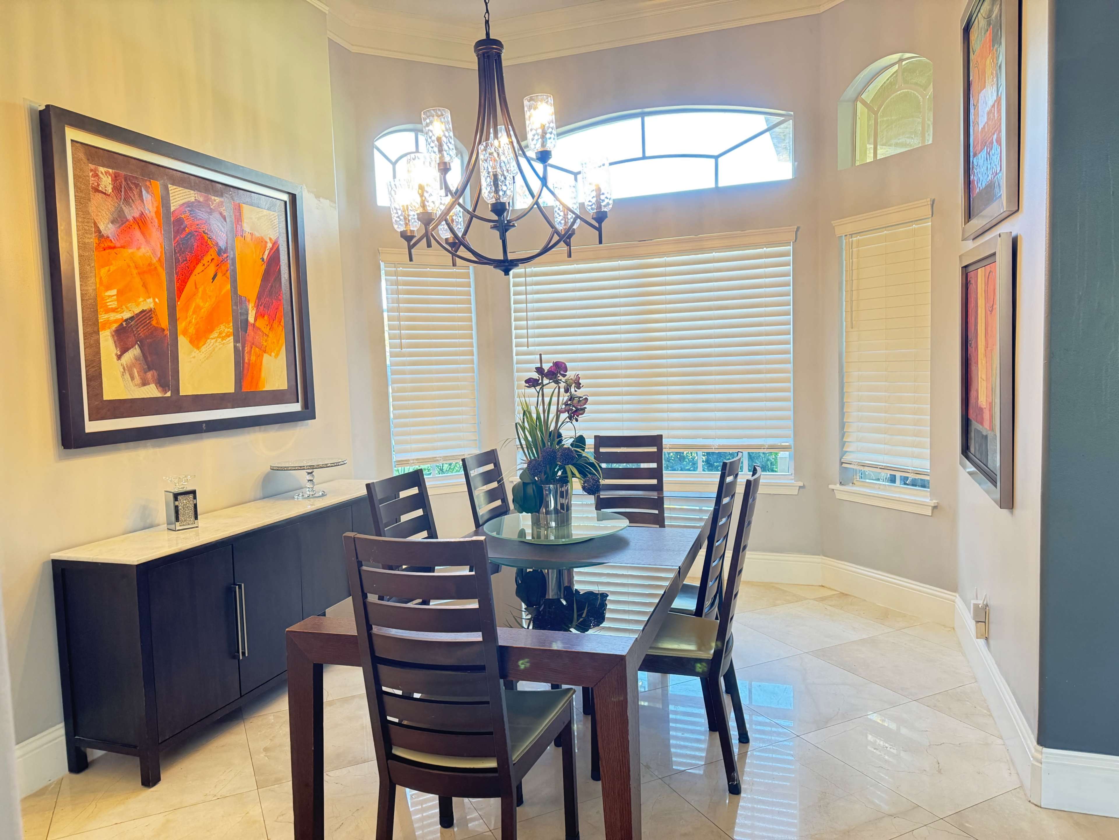 A modern dining area features a circular table surrounded by dark wooden chairs, with colorful artwork on the walls and large windows letting in natural light.