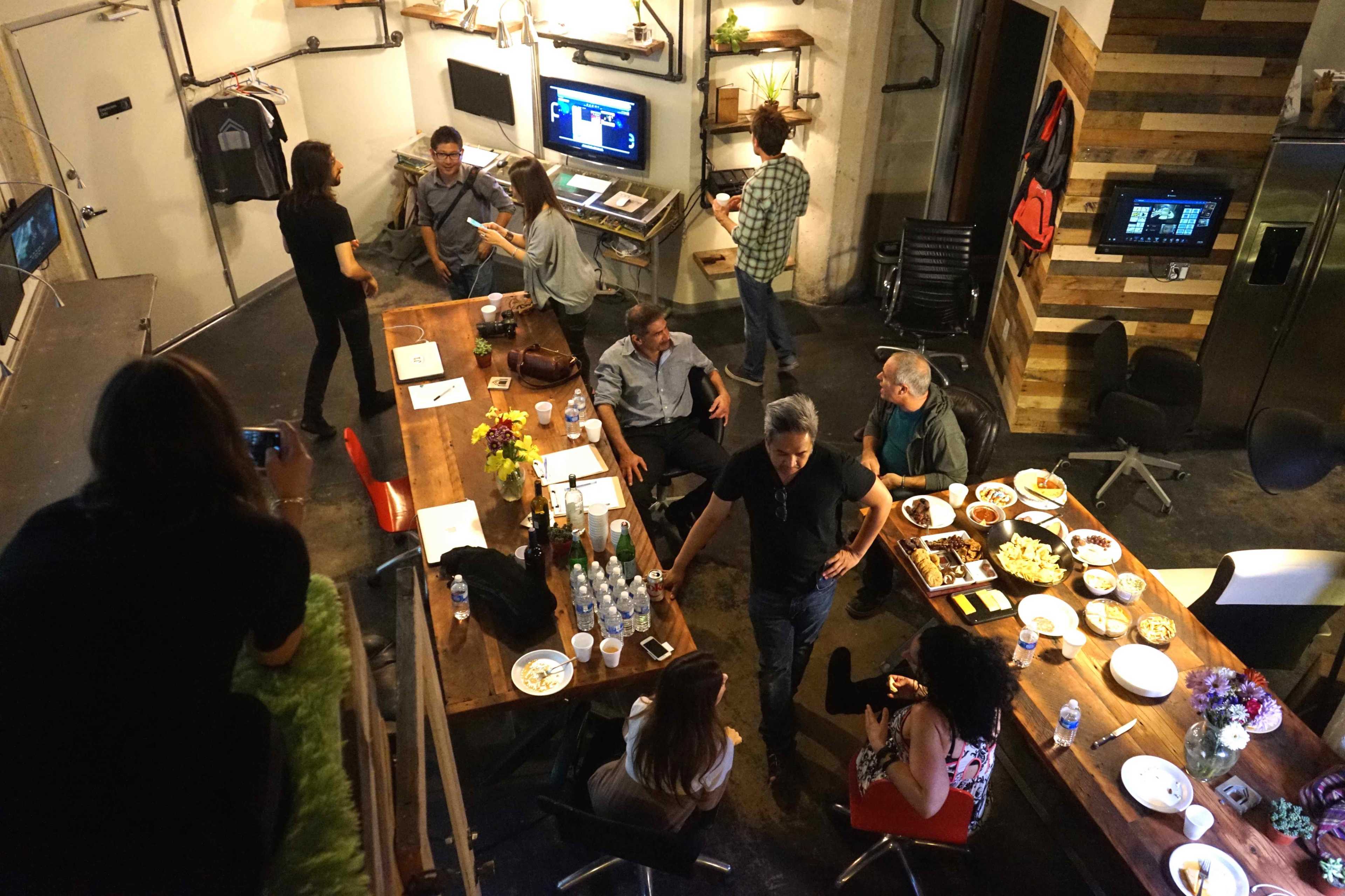 A group of people is gathered in an office-like space with food and drinks on a table, while others are engaged in conversation or working at computers.