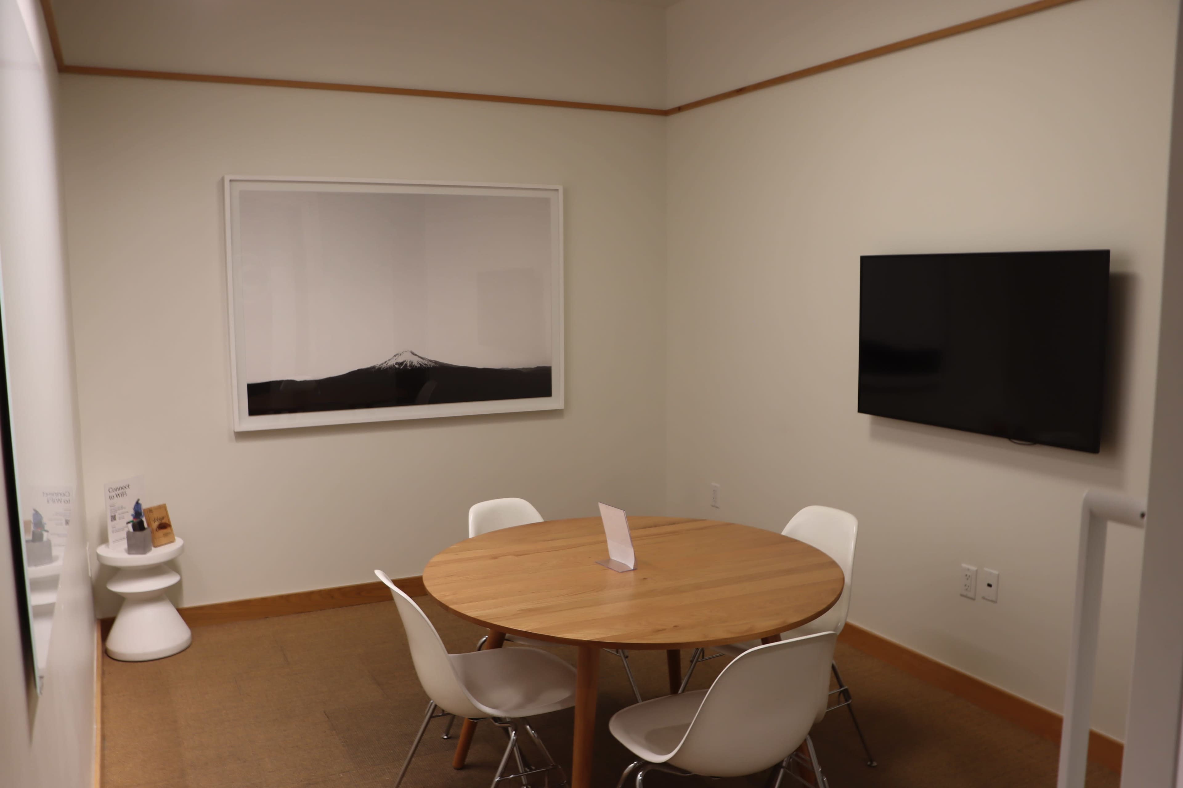 A small meeting room features a round wooden table surrounded by white chairs, with a large framed picture on the wall and a flat-screen TV mounted nearby.