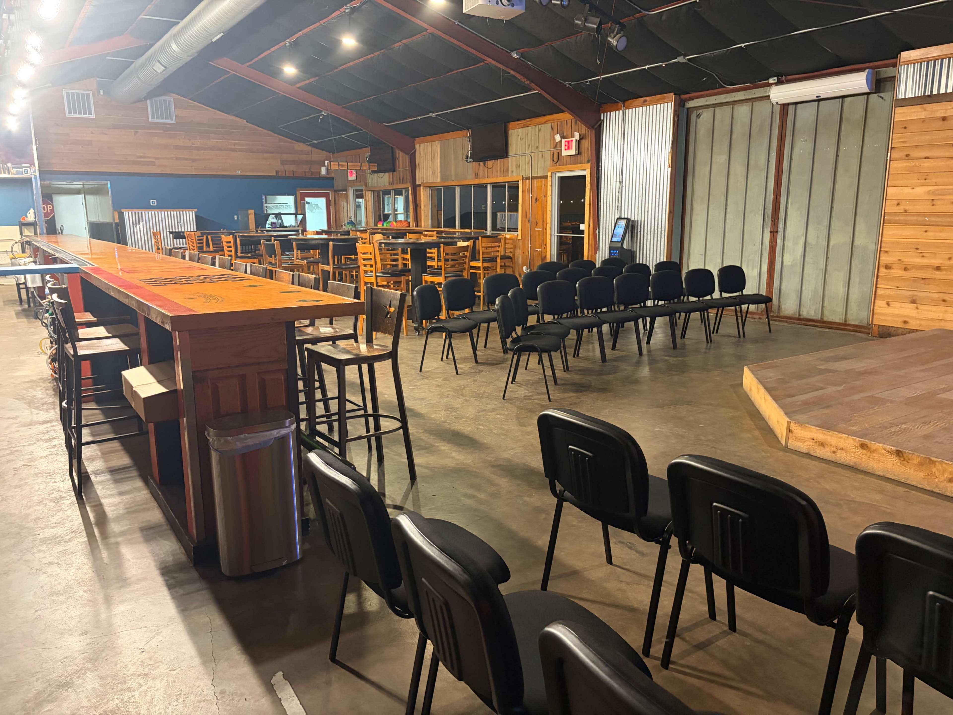 The image shows an interior venue with a wooden bar on the left, empty chairs arranged in rows facing a small stage, and a casual dining area in the background.