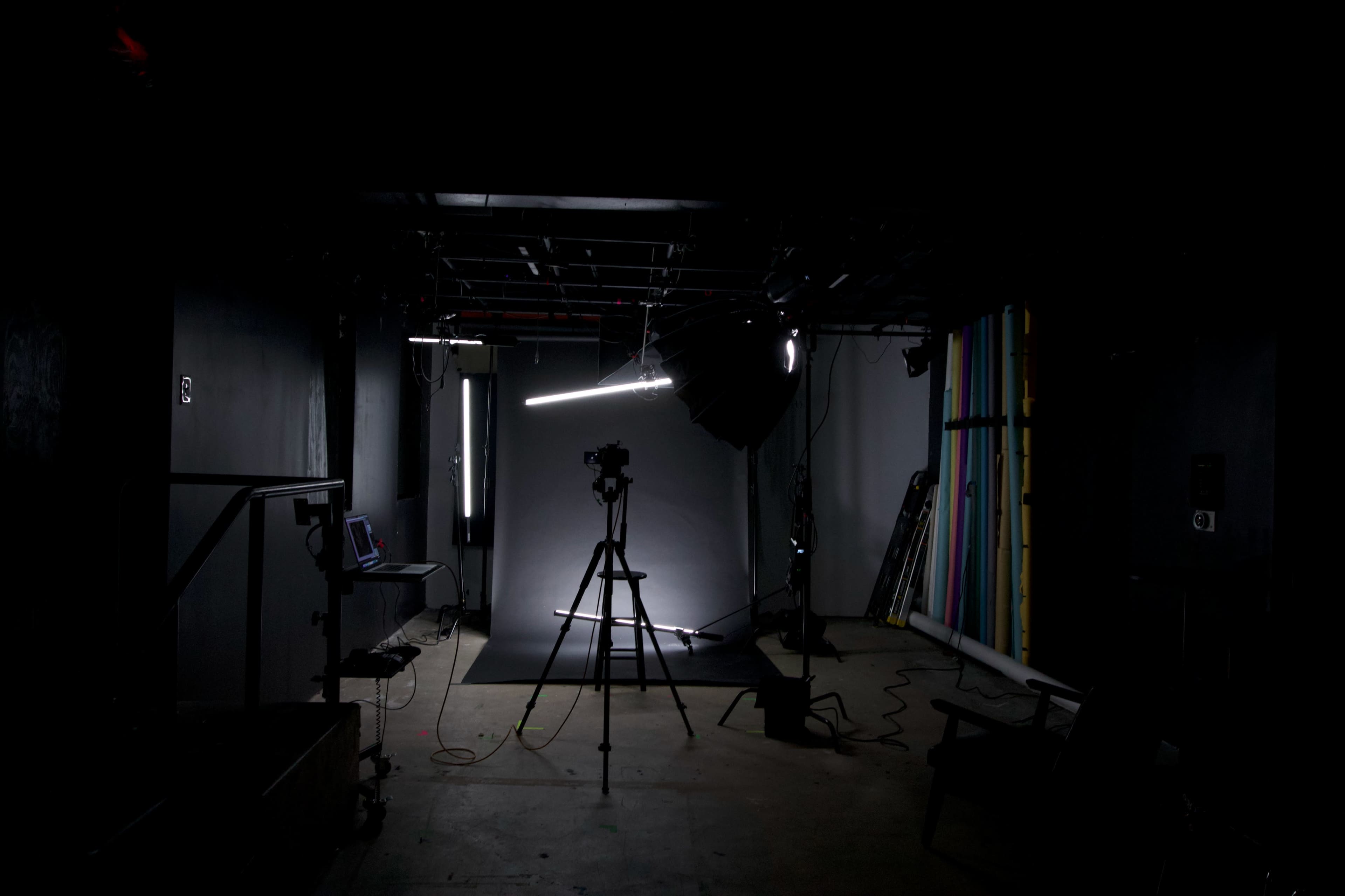 A dimly lit photography studio is set up with a tripod, camera, and backdrop, surrounded by various studio equipment and lighting.