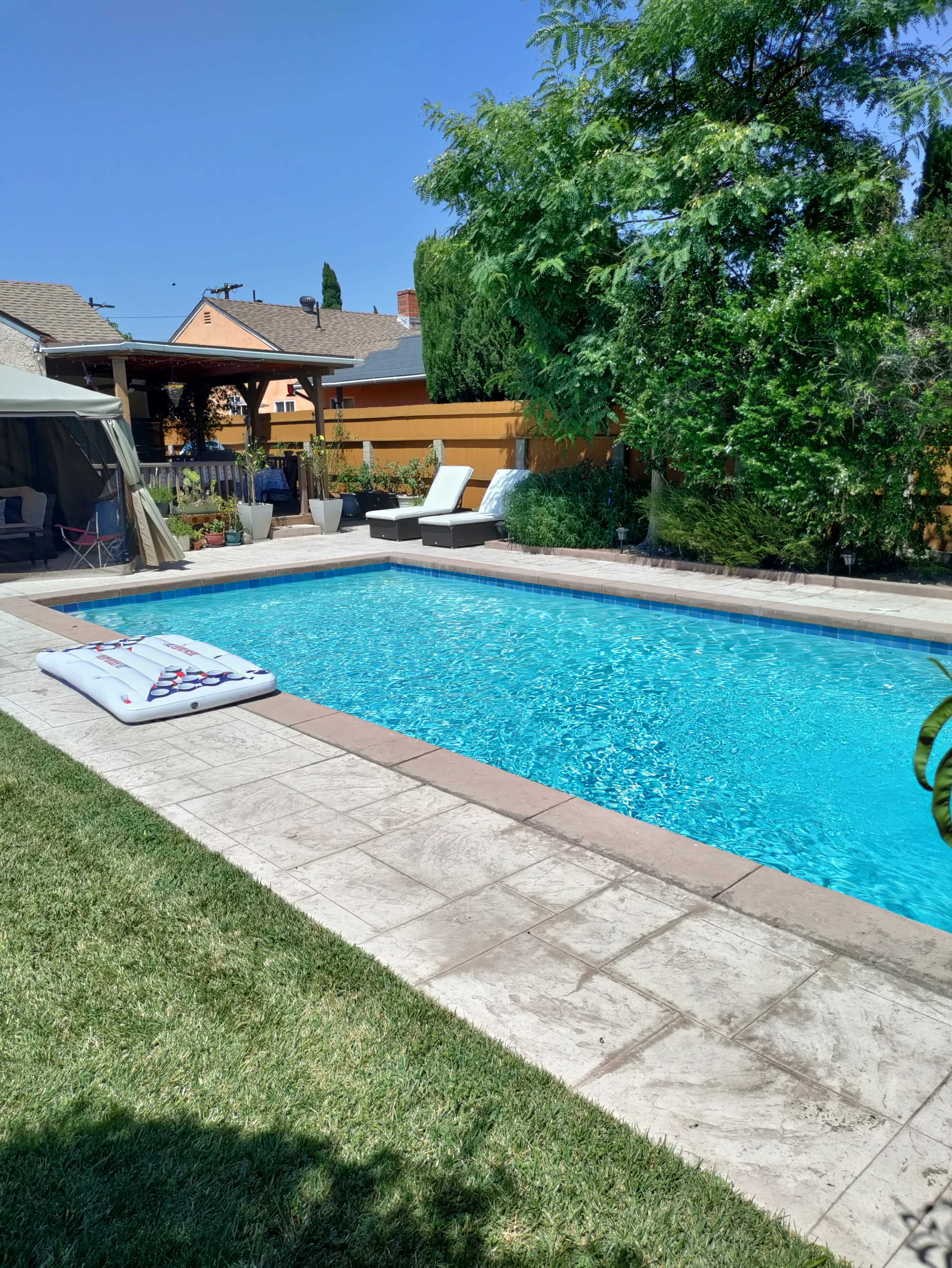 The image shows a clear swimming pool surrounded by a patio, lounge chairs, and greenery in a residential backyard.