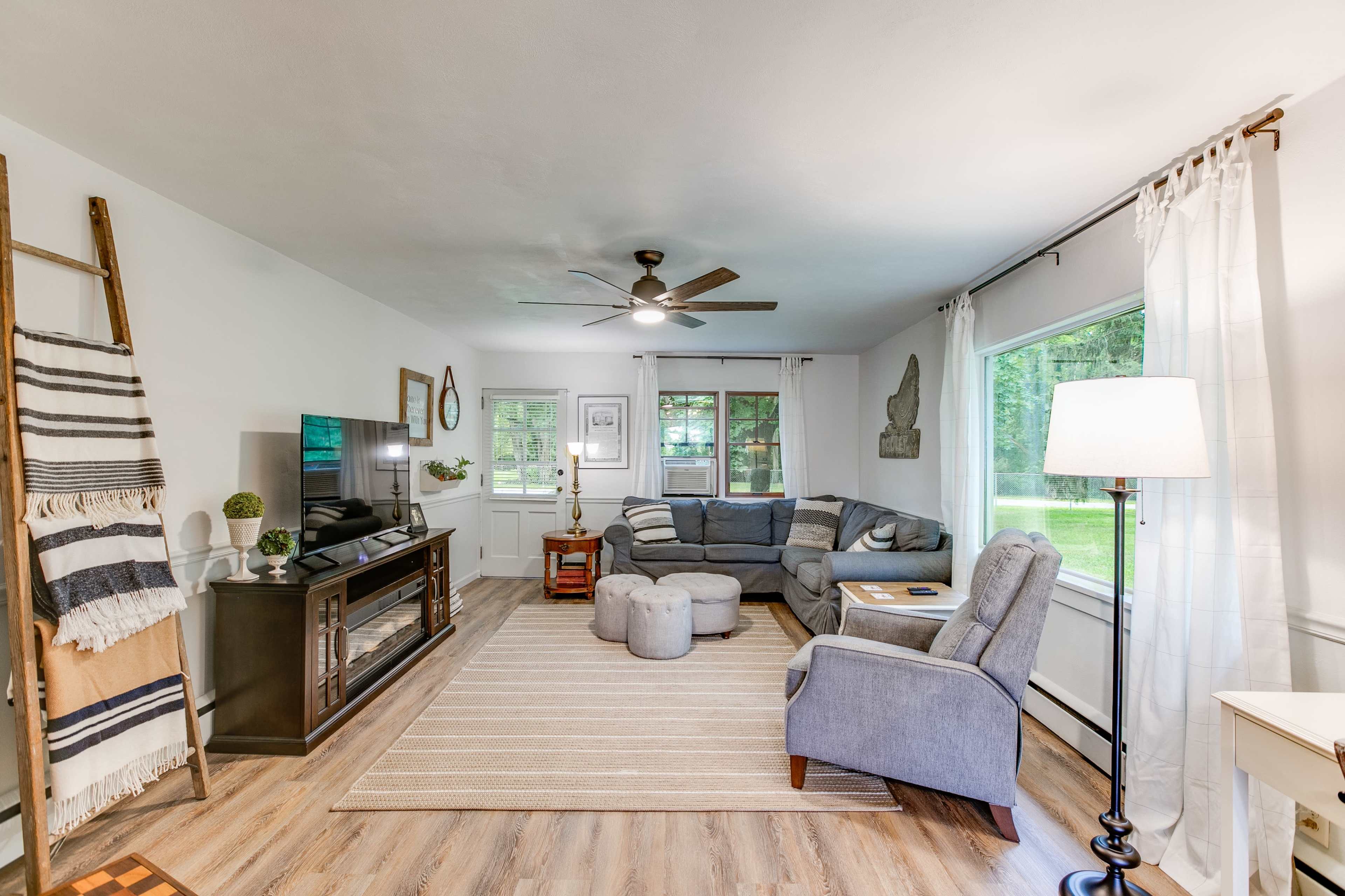 A living room with a gray sofa, a light rug, a television stand, and large windows allowing natural light to enter.