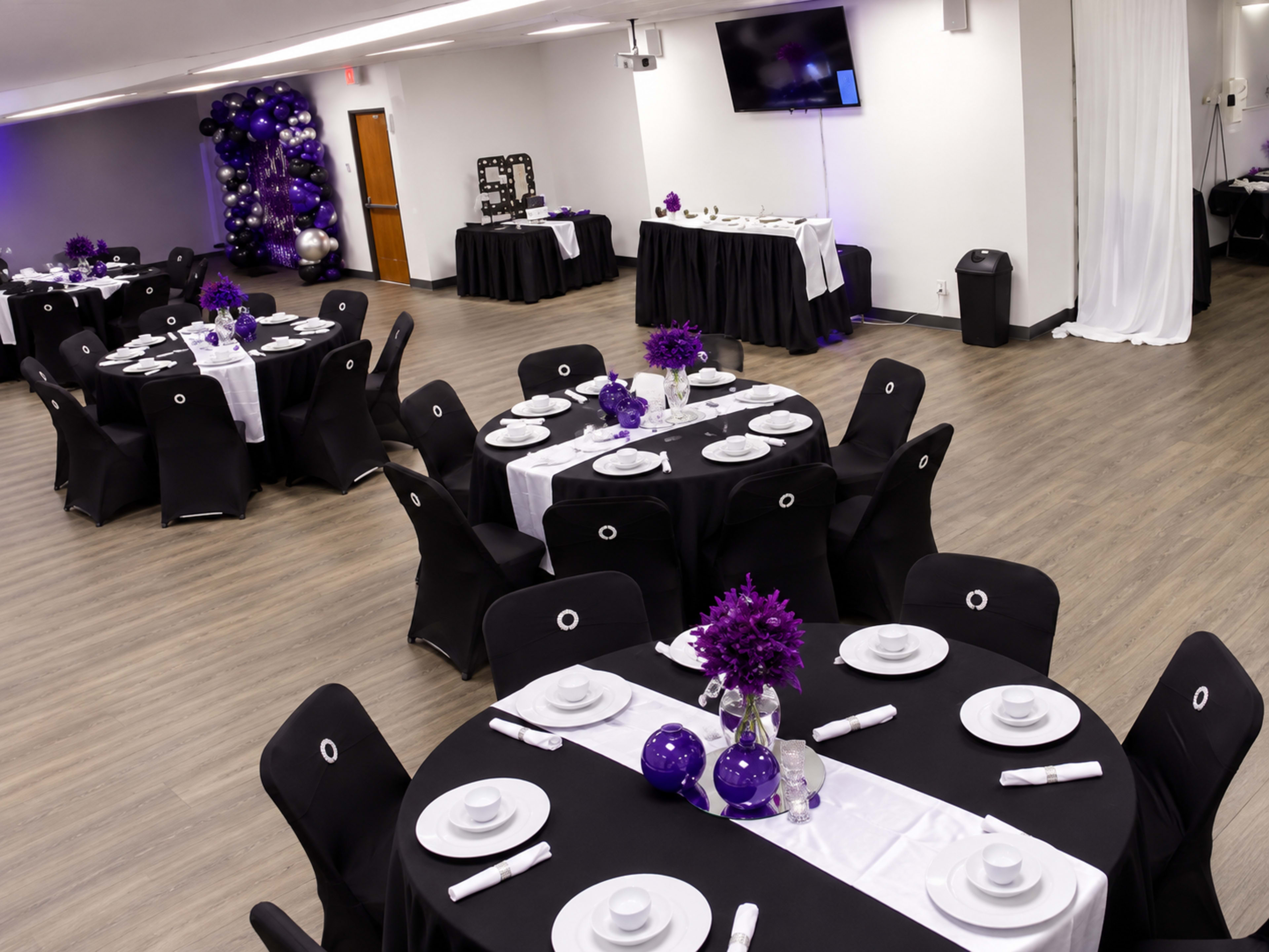 The image shows a spacious event room set up with neatly arranged black tables, each featuring white tableware and purple floral centerpieces.