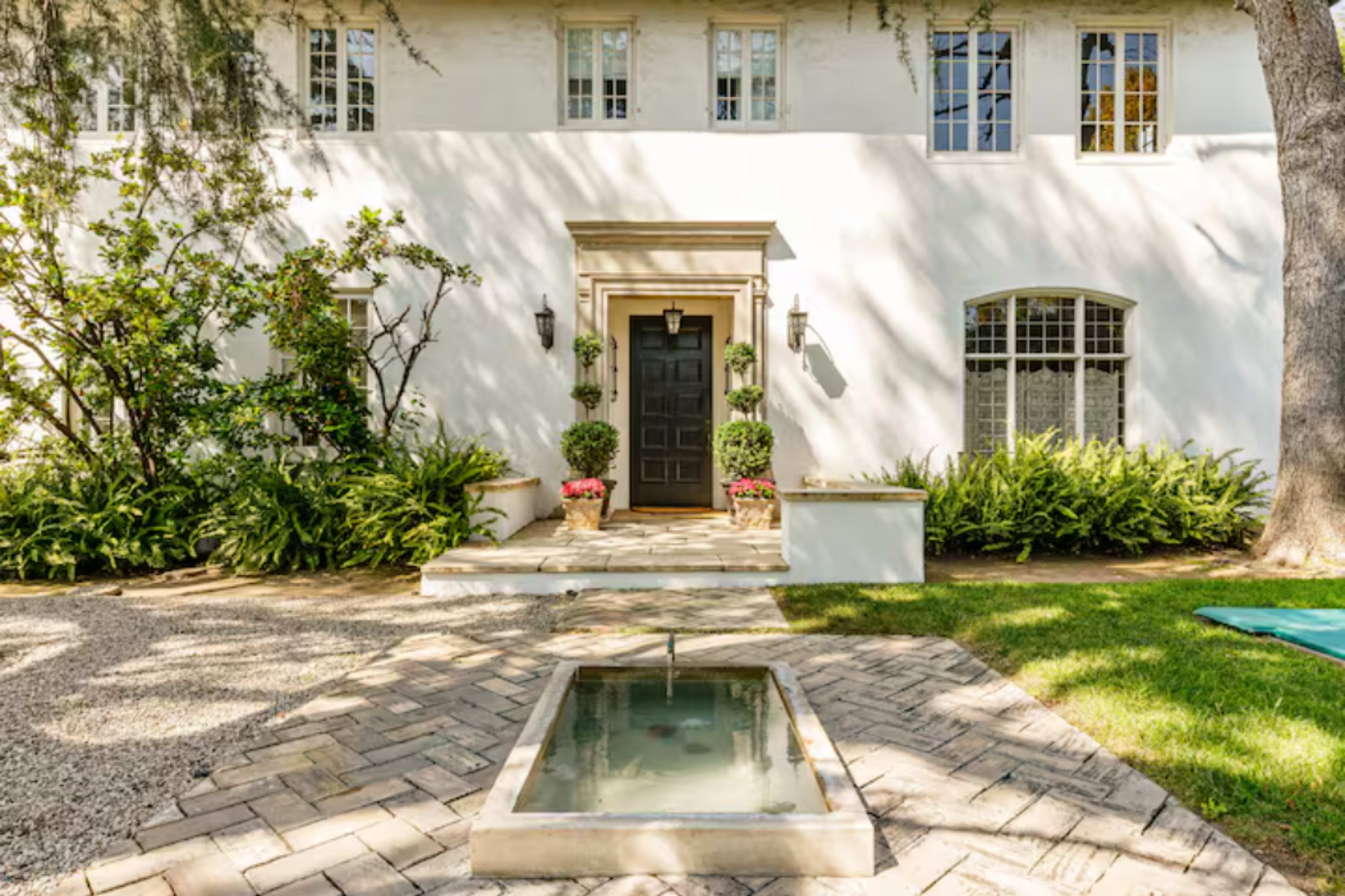 A well-maintained white house features a central black door, flanked by potted plants and a small fountain in the foreground.