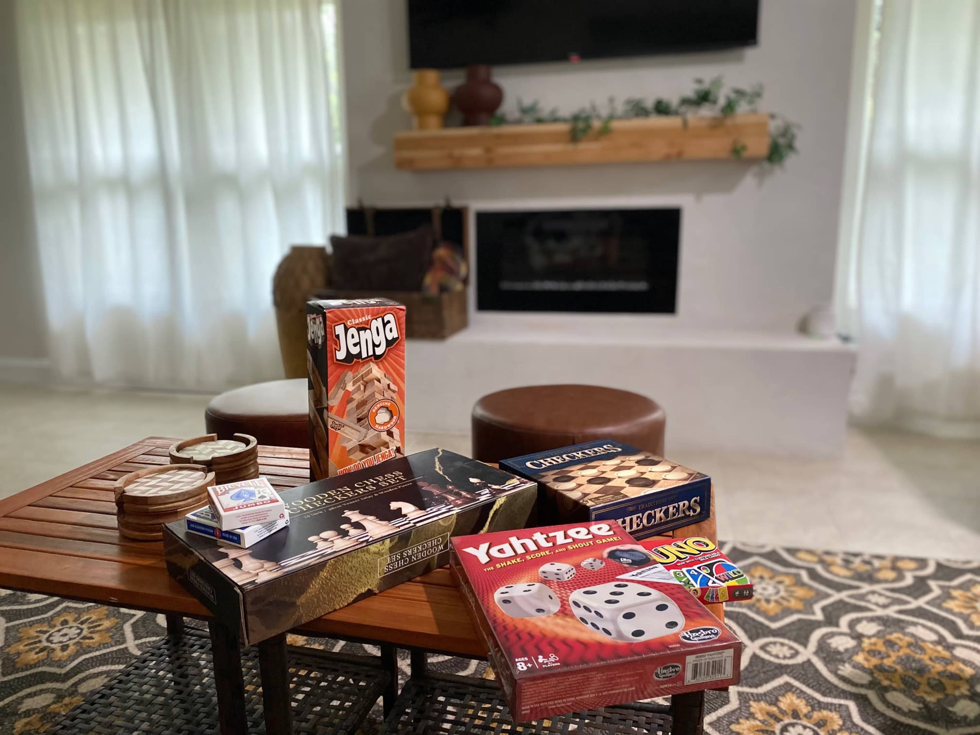 A wooden coffee table displays various board games, including Jenga, Uno, and Yahtzee, in a living room with a fireplace and a large window.