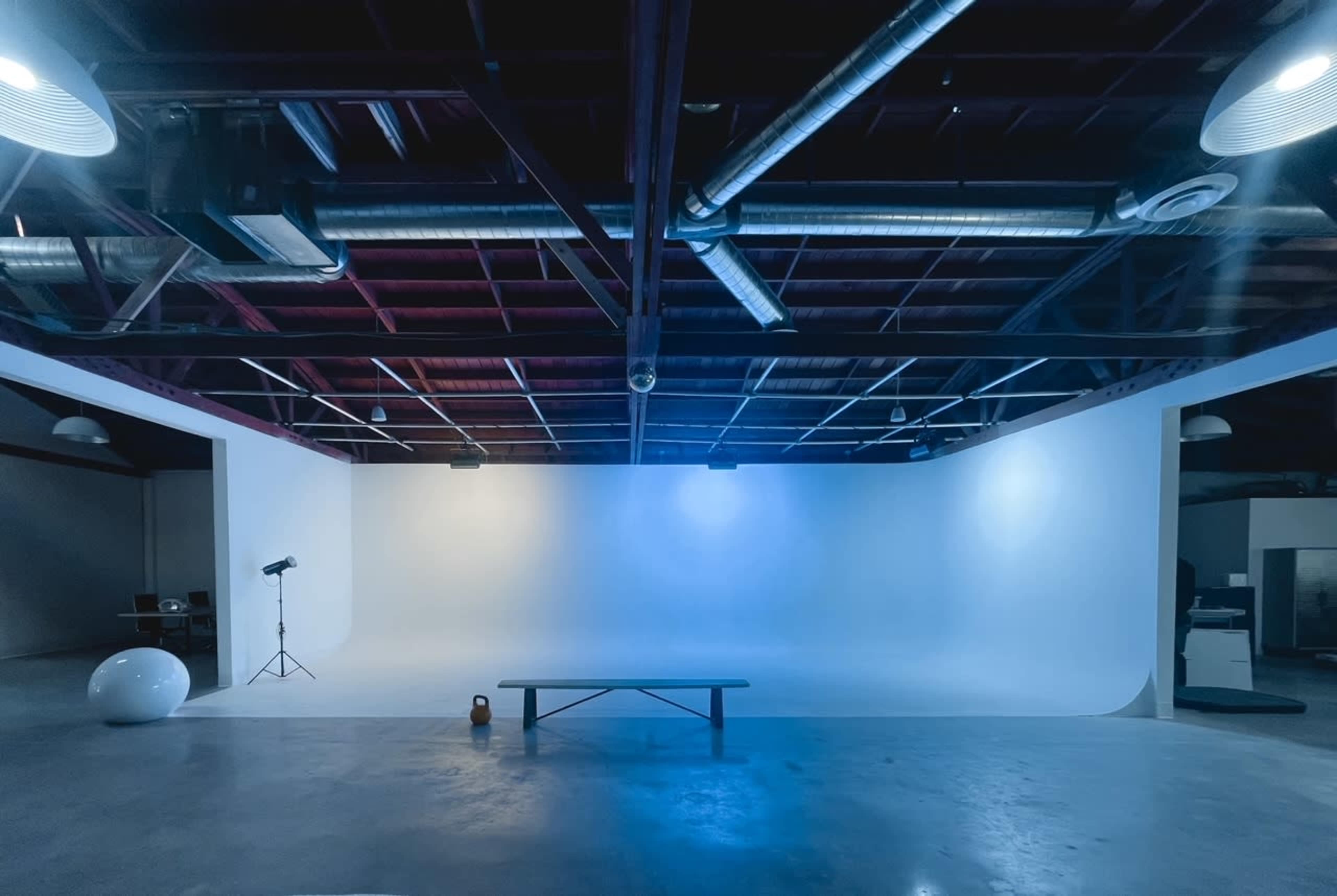 An empty studio space features a smooth white backdrop, a wooden bench, a round object, and various lighting equipment suspended from the ceiling.