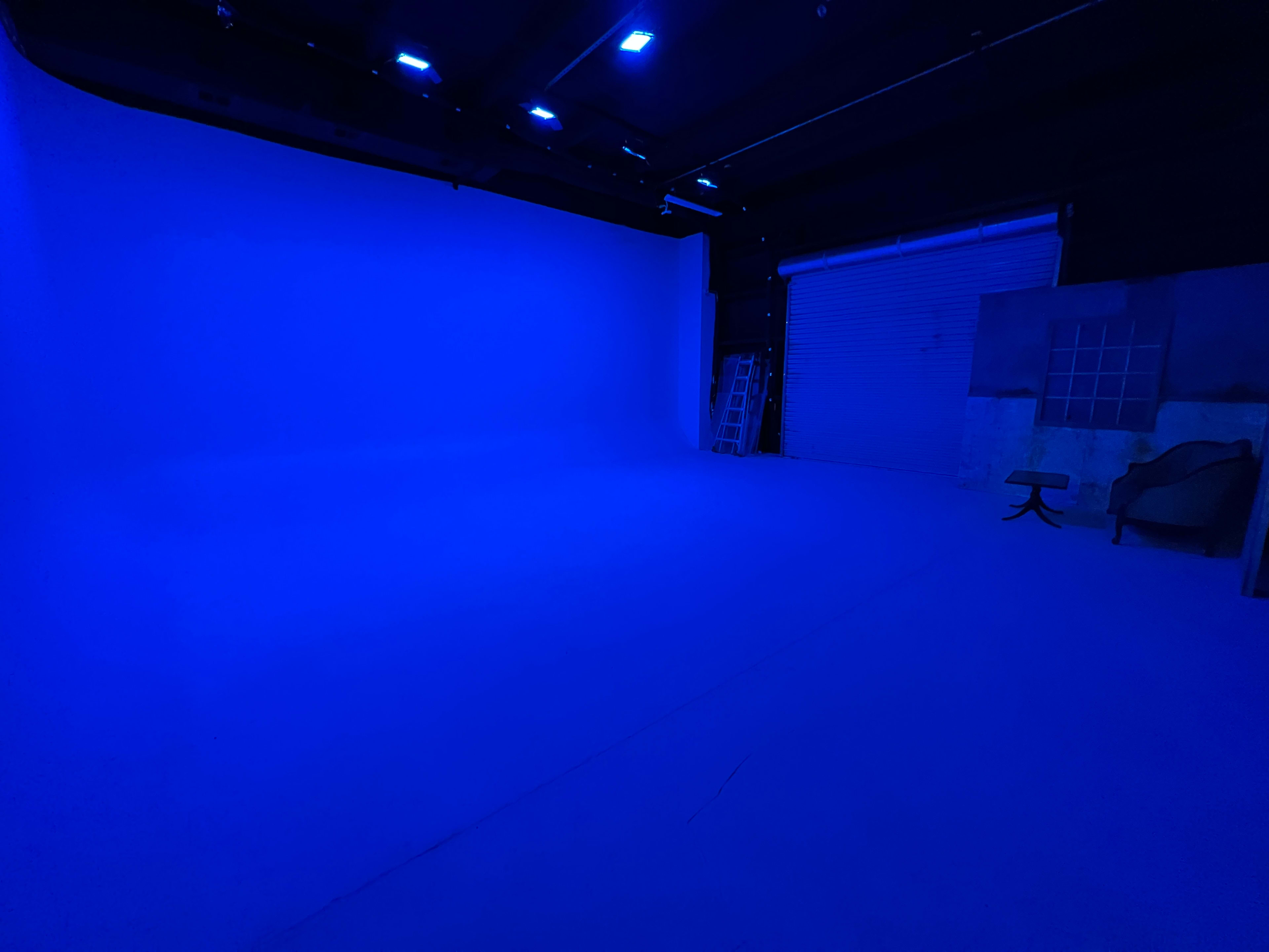 A large studio space is illuminated with blue light, featuring a smooth floor and a backdrop of a solid blue wall, along with a ladder and a small table in the corner.