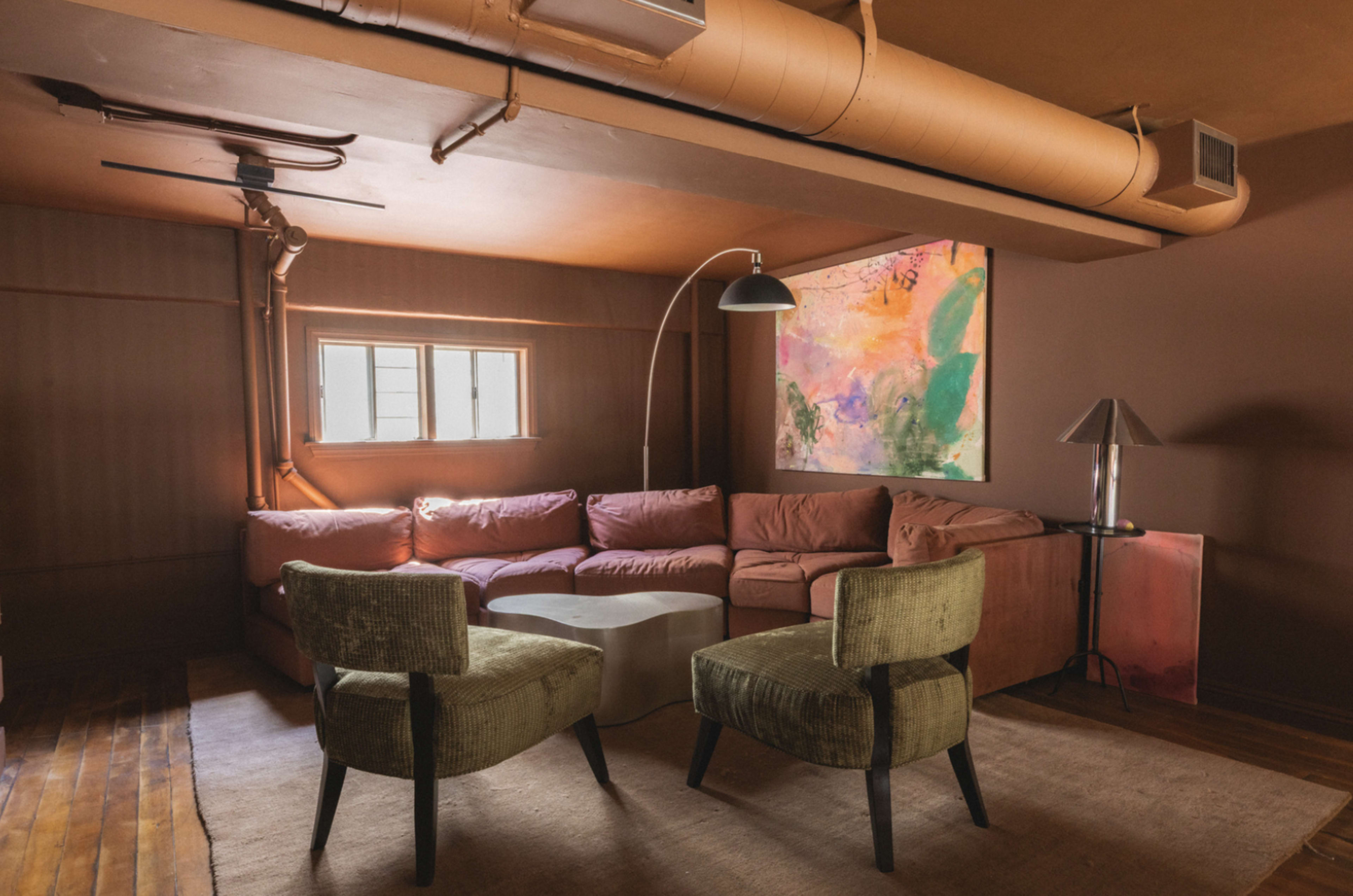 The image shows a cozy living room with a pink sectional sofa, two green armchairs, and a large abstract painting on the wall.
