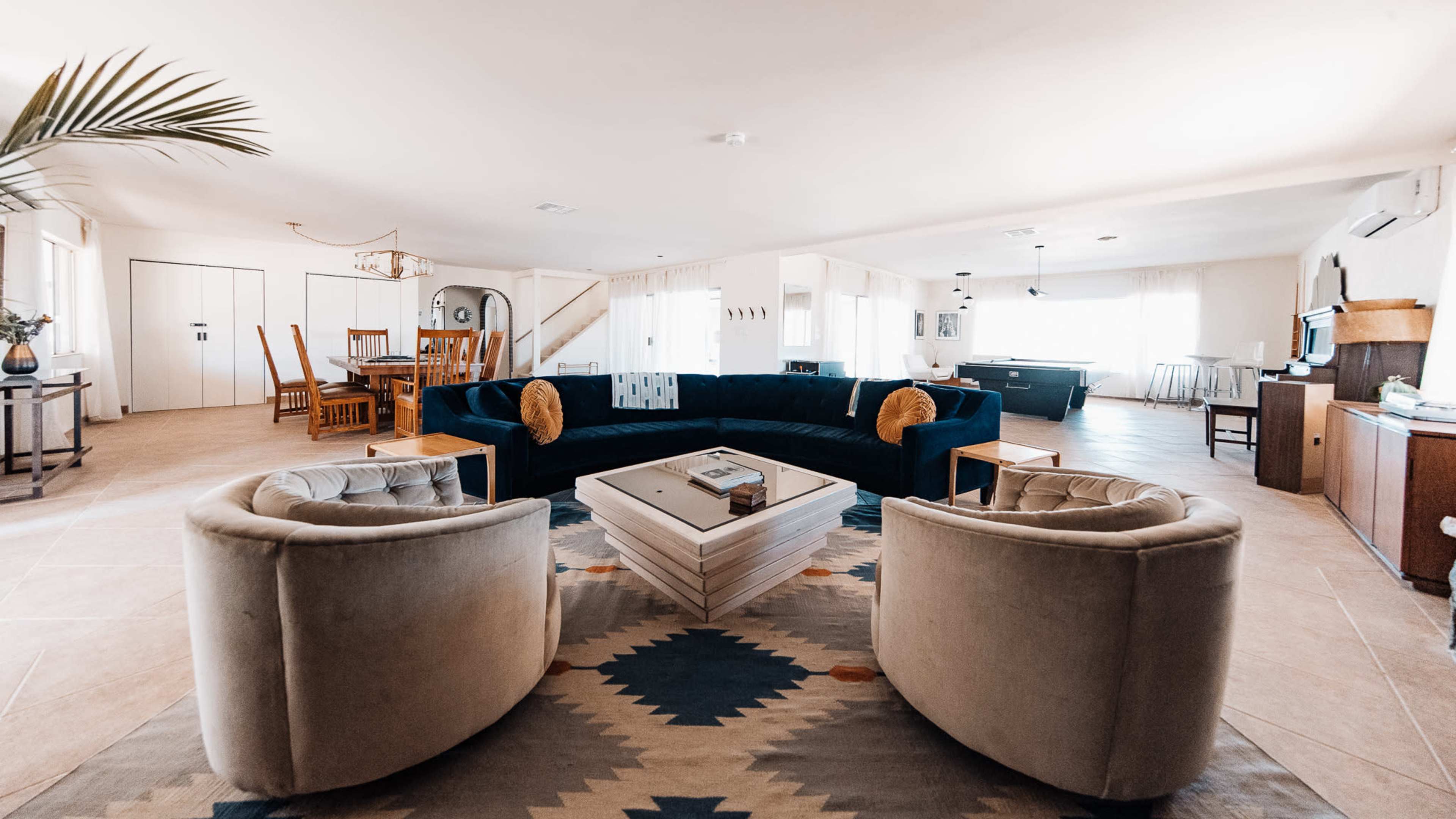 The image shows a spacious living area featuring a large blue sectional sofa, two circular chairs, a white coffee table, and a dining area in the background.