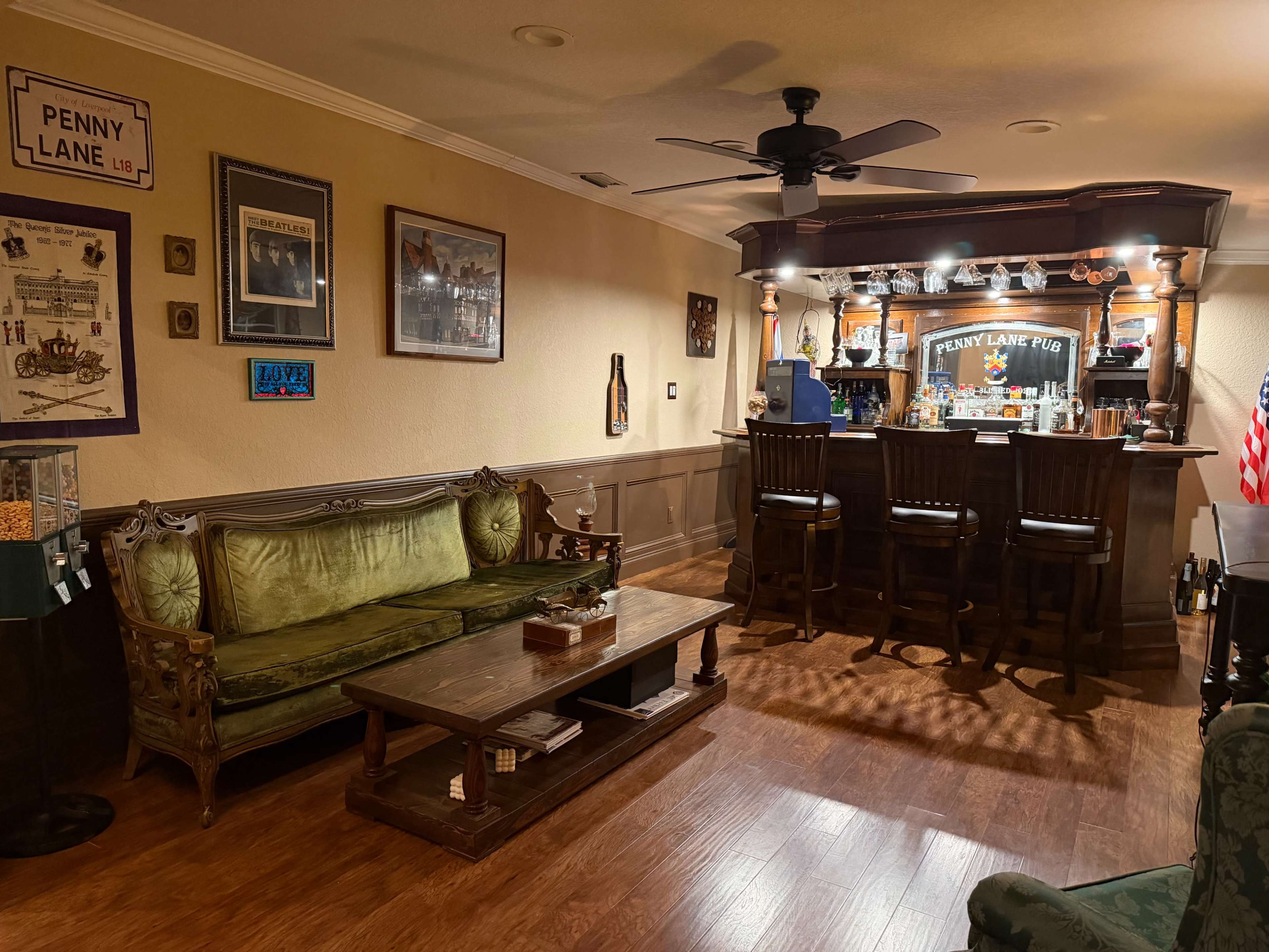 The image shows a cozy home bar area with a wooden bar counter, several stools, a green velvet couch, and a coffee table, all illuminated by warm lights.