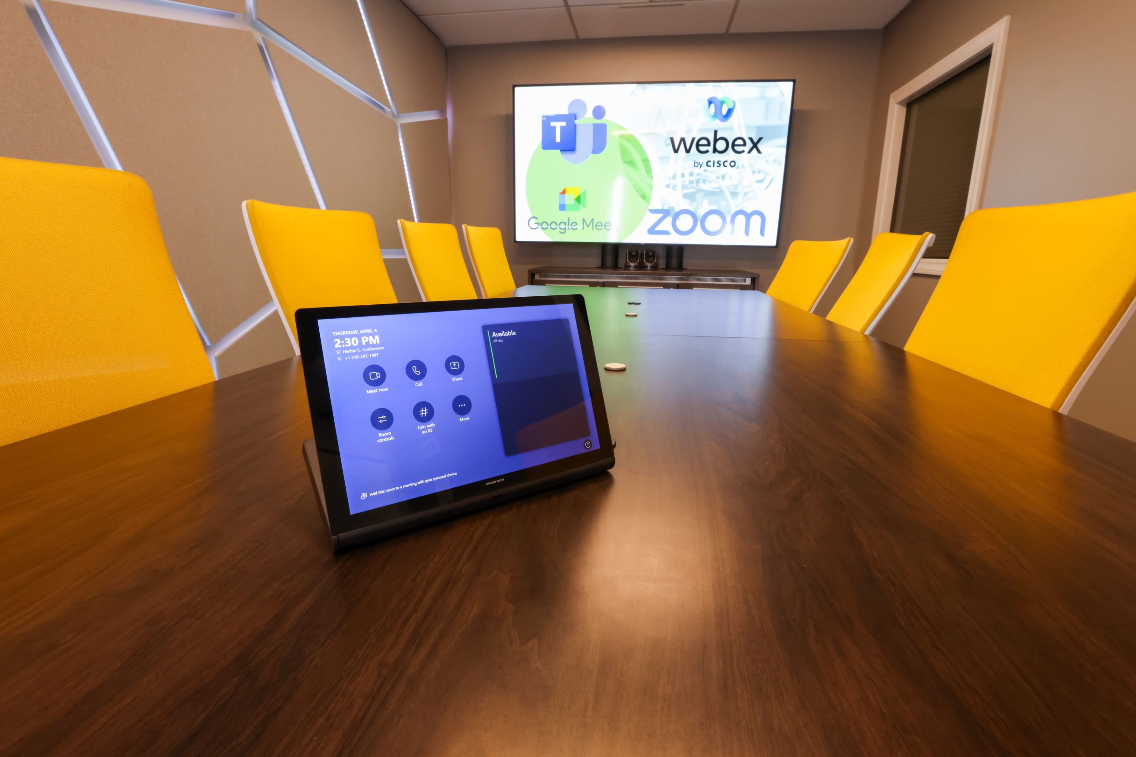 A conference room with a wooden table, a tablet displaying a meeting interface, and a large screen showing video conferencing logos.