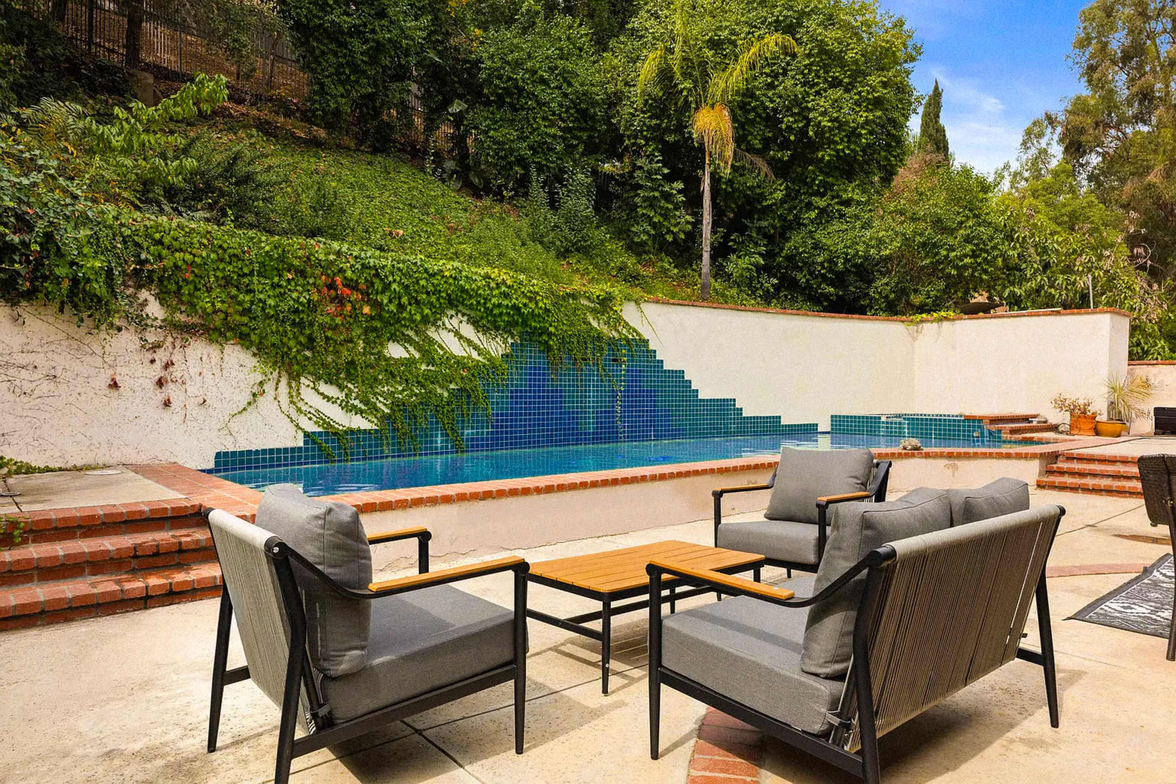 The image shows a patio area with two lounge chairs and a coffee table overlooking a tiled swimming pool surrounded by greenery.