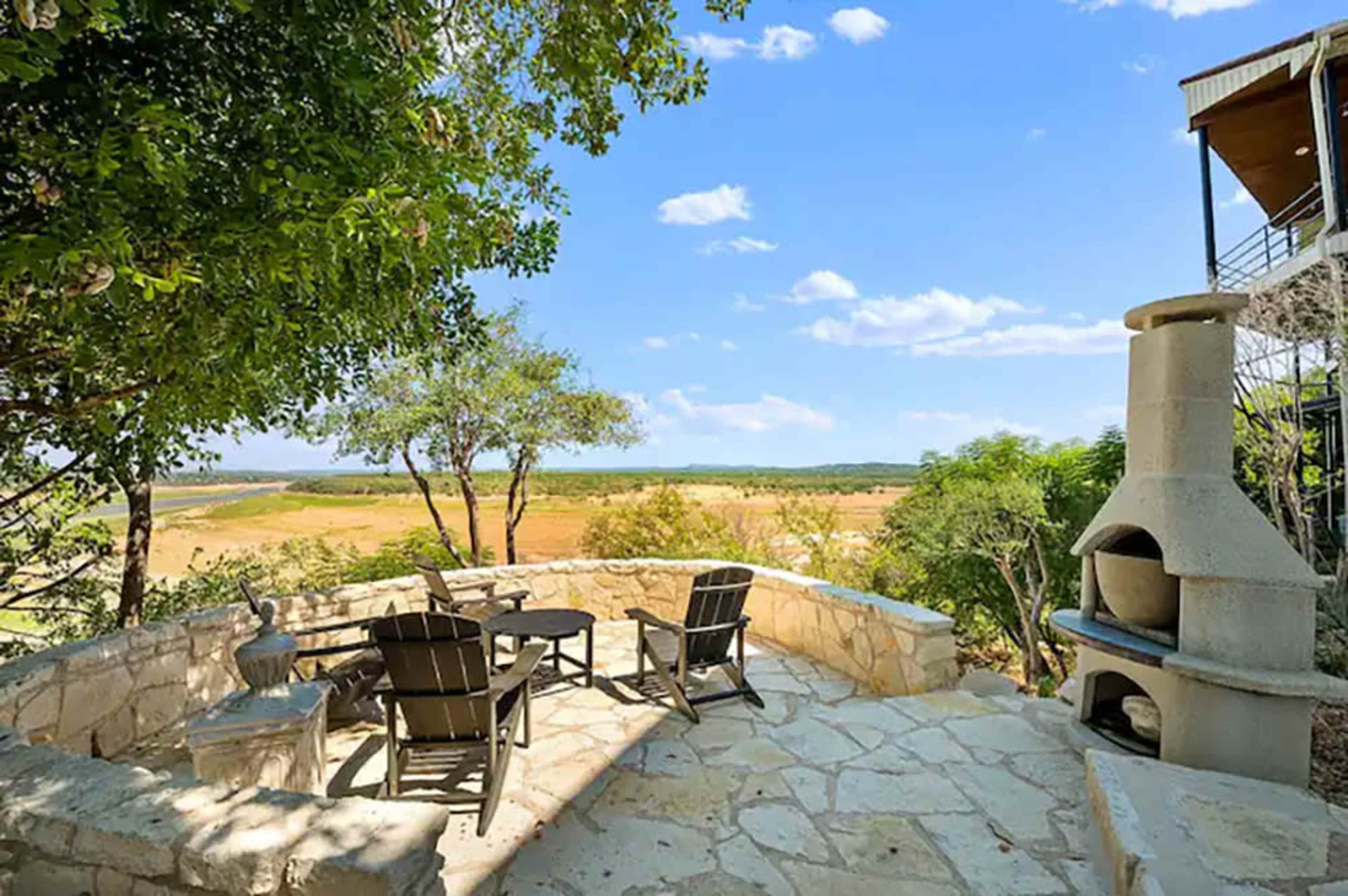 A stone patio with outdoor seating and a stone fireplace, overlooking a grassy landscape under a clear blue sky.