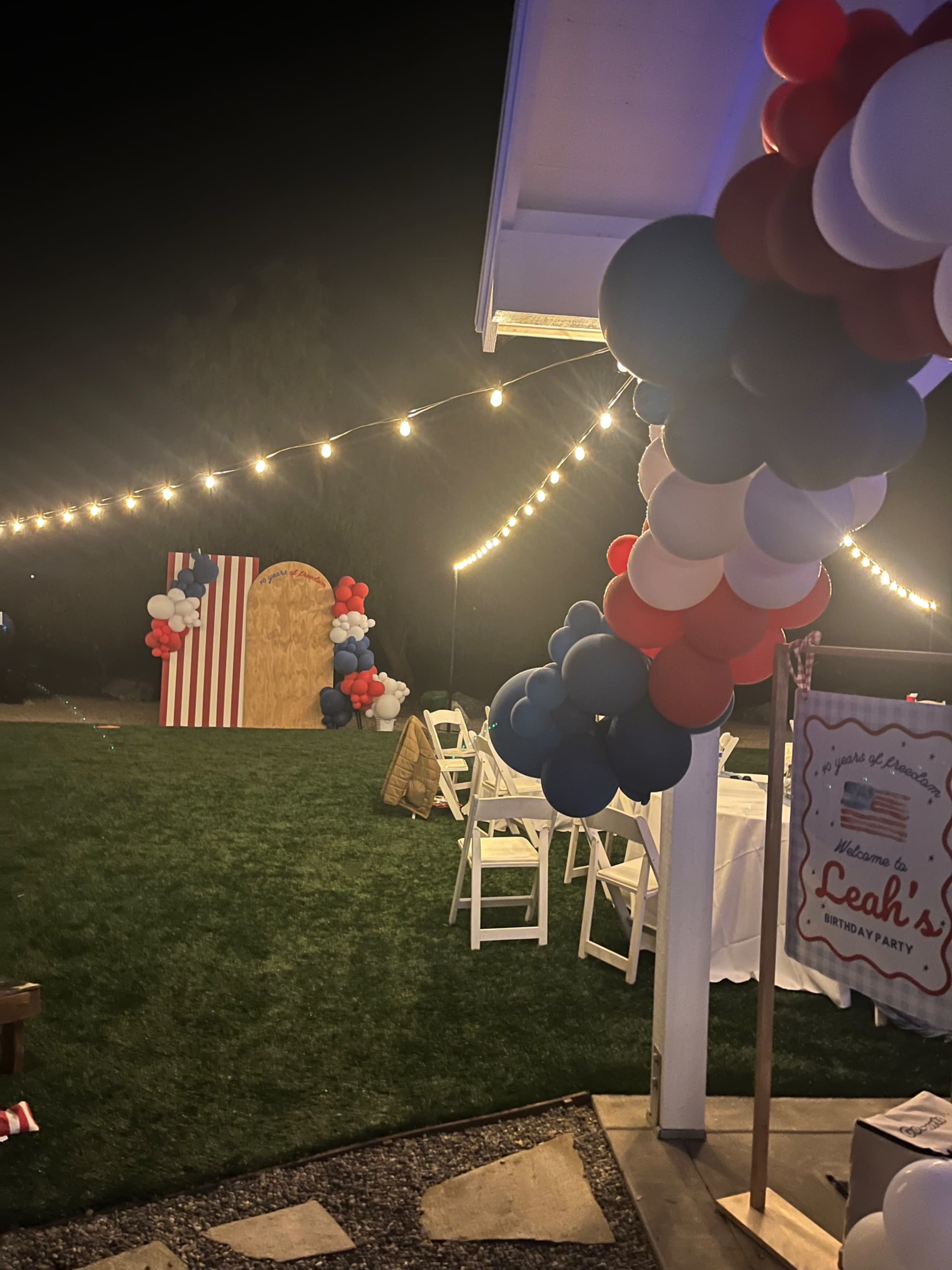 The image shows a decorated outdoor party setup with string lights, balloons in red, white, and blue, and a welcome sign for a birthday celebration.