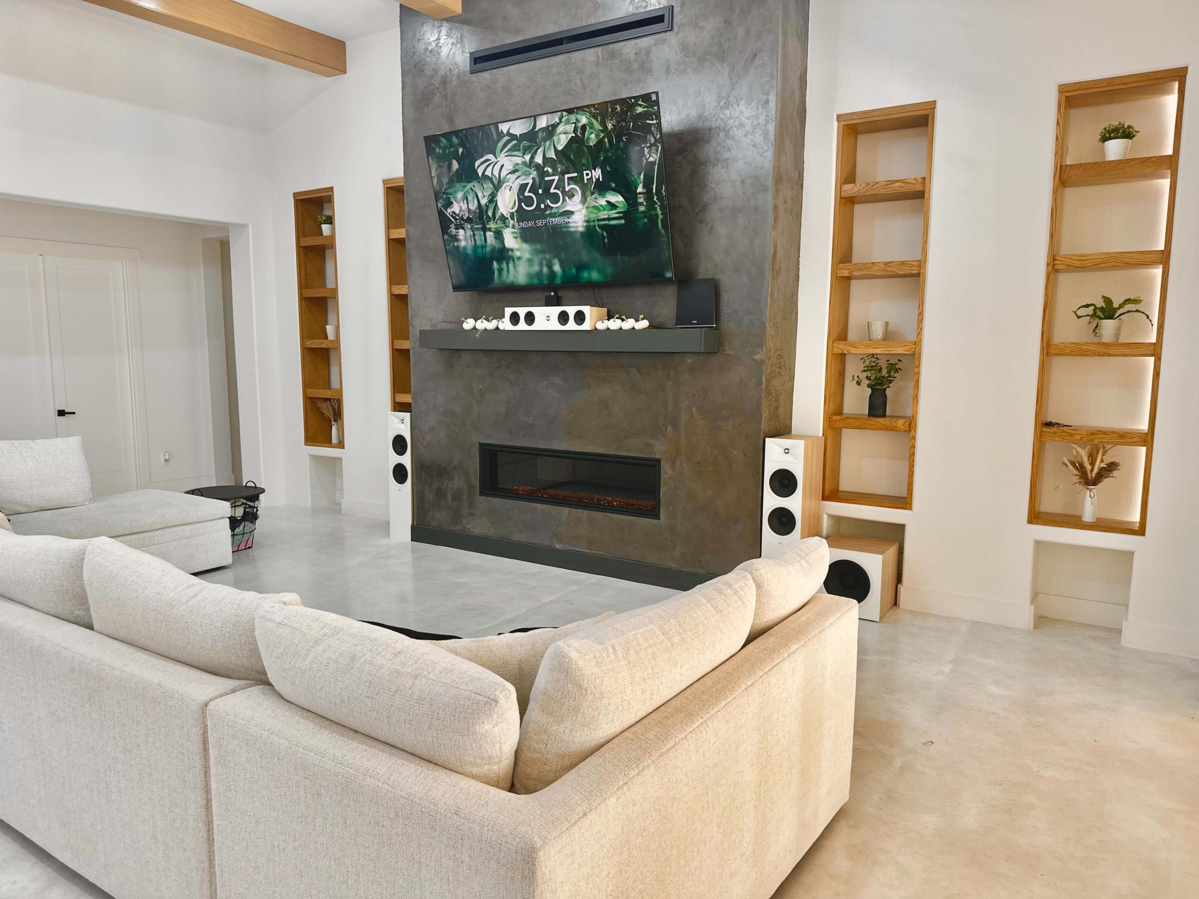 A modern living room features a large sectional sofa facing a mounted television above a sleek fireplace, with built-in shelves on either side displaying decorative plants.