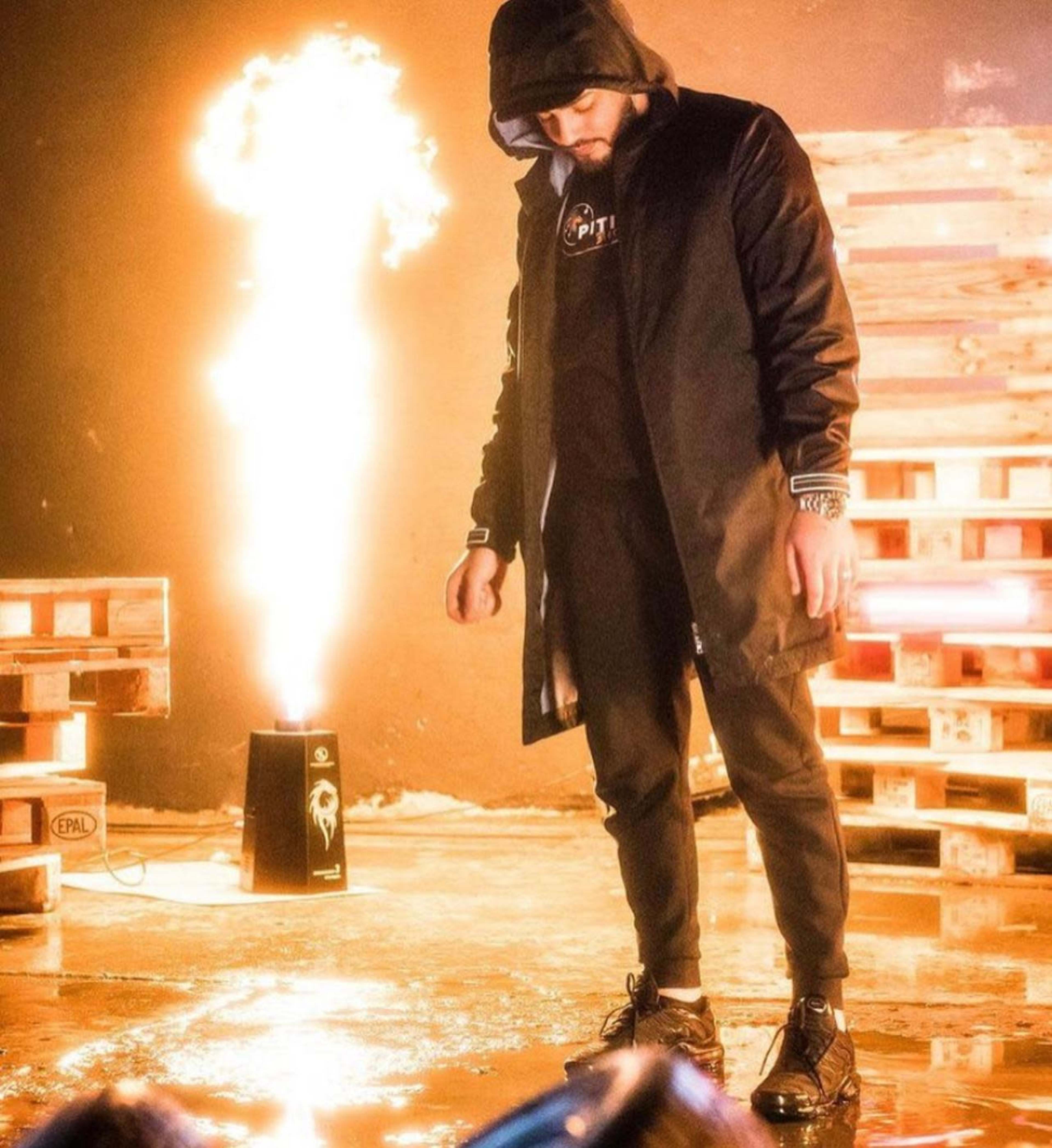 A man in a black hooded jacket and pants stands on a reflective surface in front of a flame shooting up from a speaker.