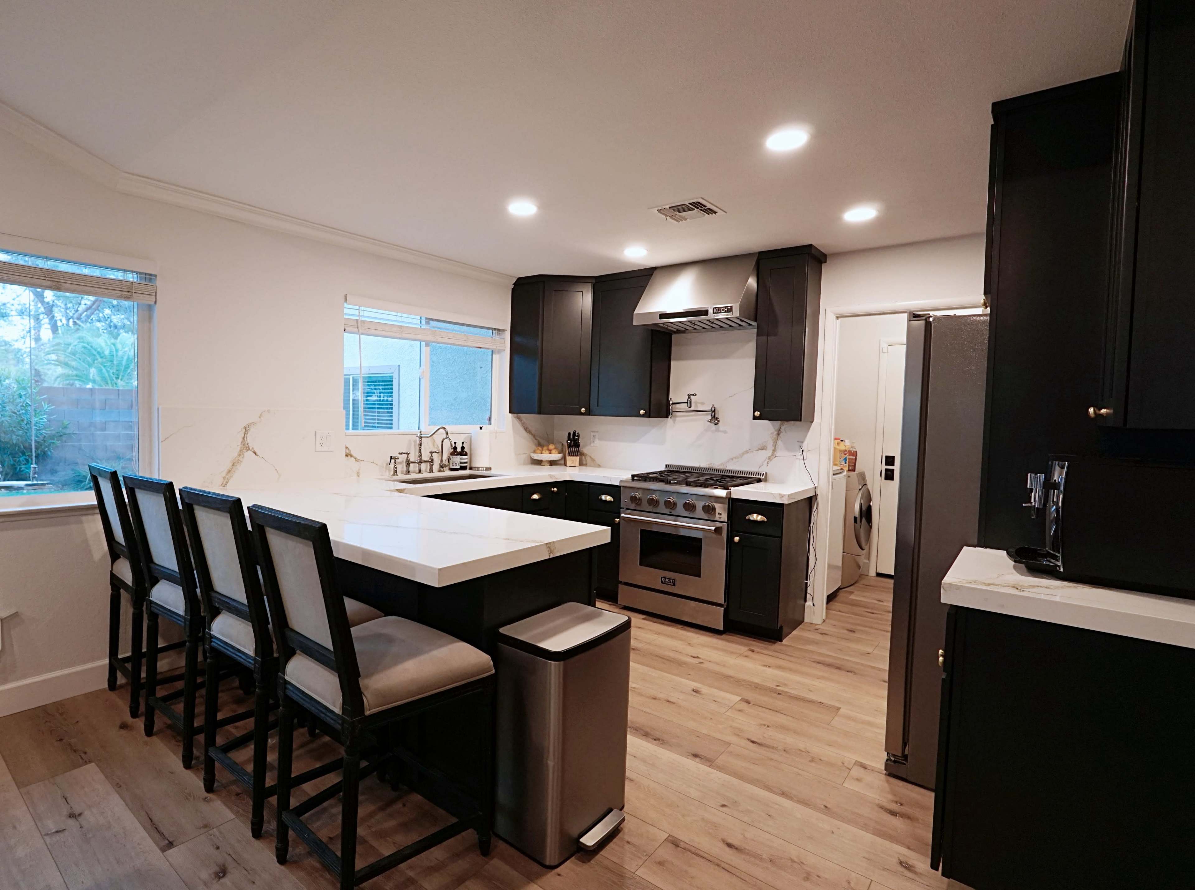 A modern kitchen features dark cabinetry, a large island with seating, stainless steel appliances, and ample natural light from windows.