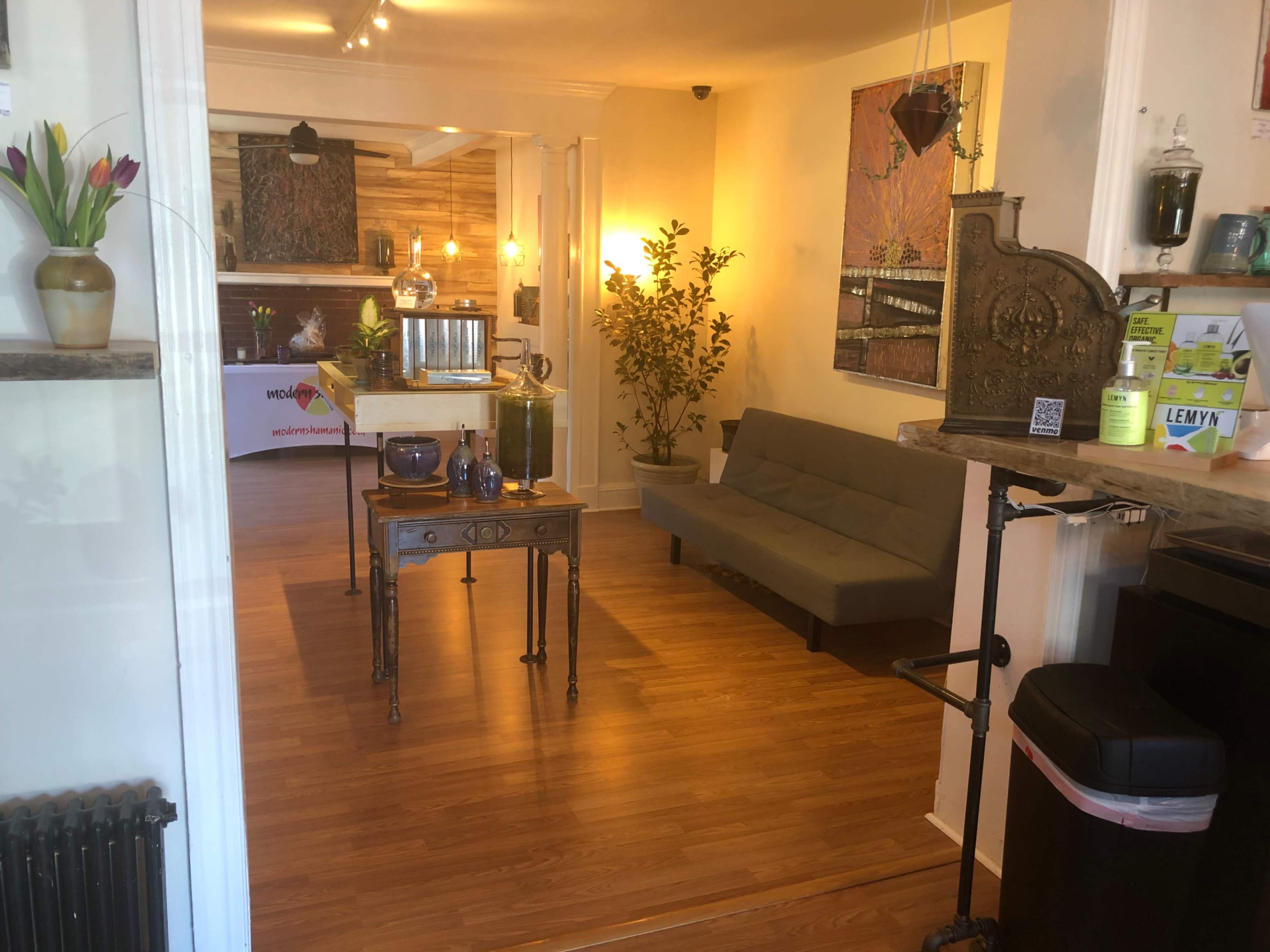 A modern reception area with a sofa, a table set with items, and decorative plants, leading to a kitchen or dining space in the background.