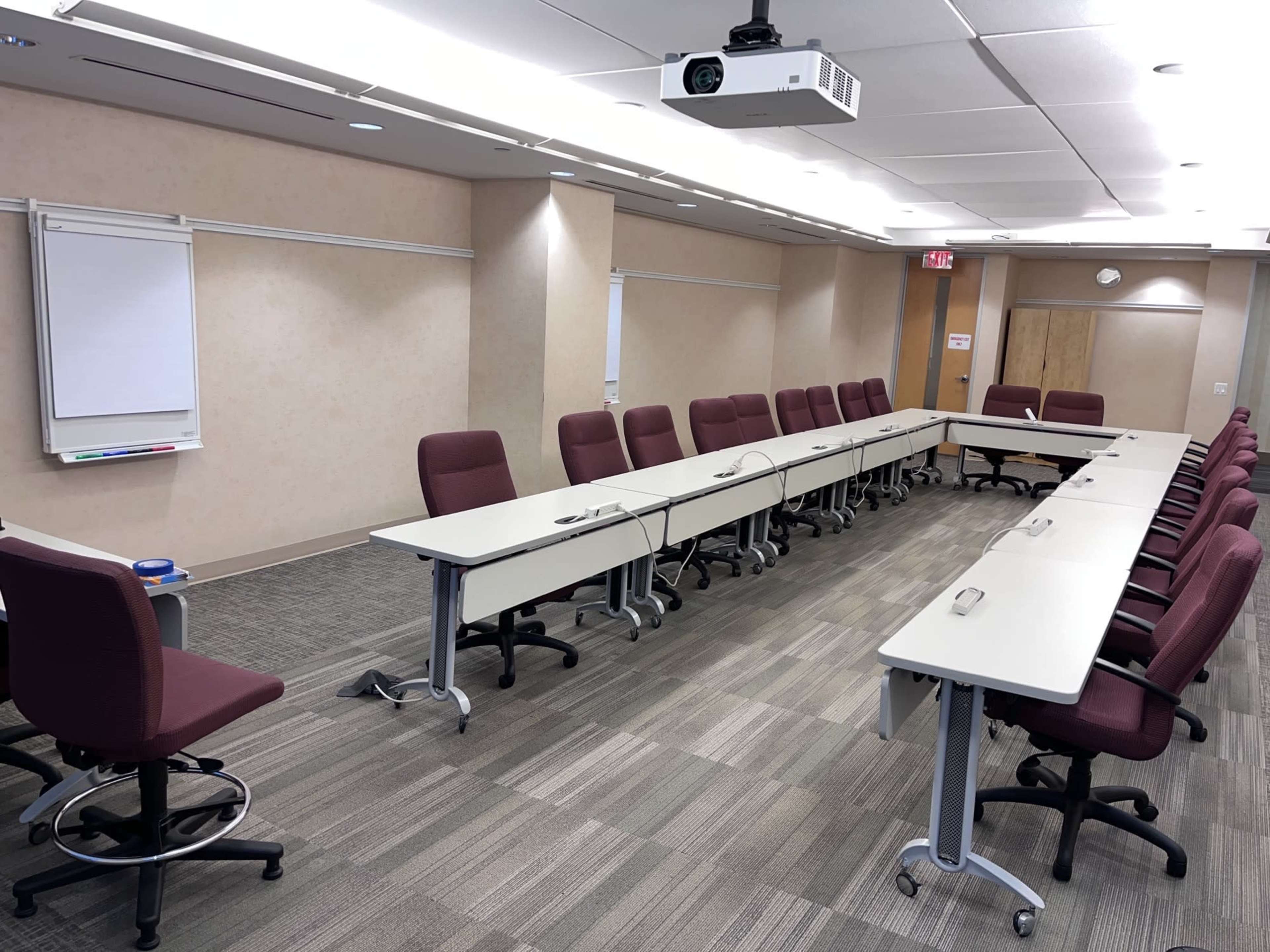 The image shows a conference room with several rows of tables and chairs arranged for a meeting, featuring a projector mounted on the ceiling.