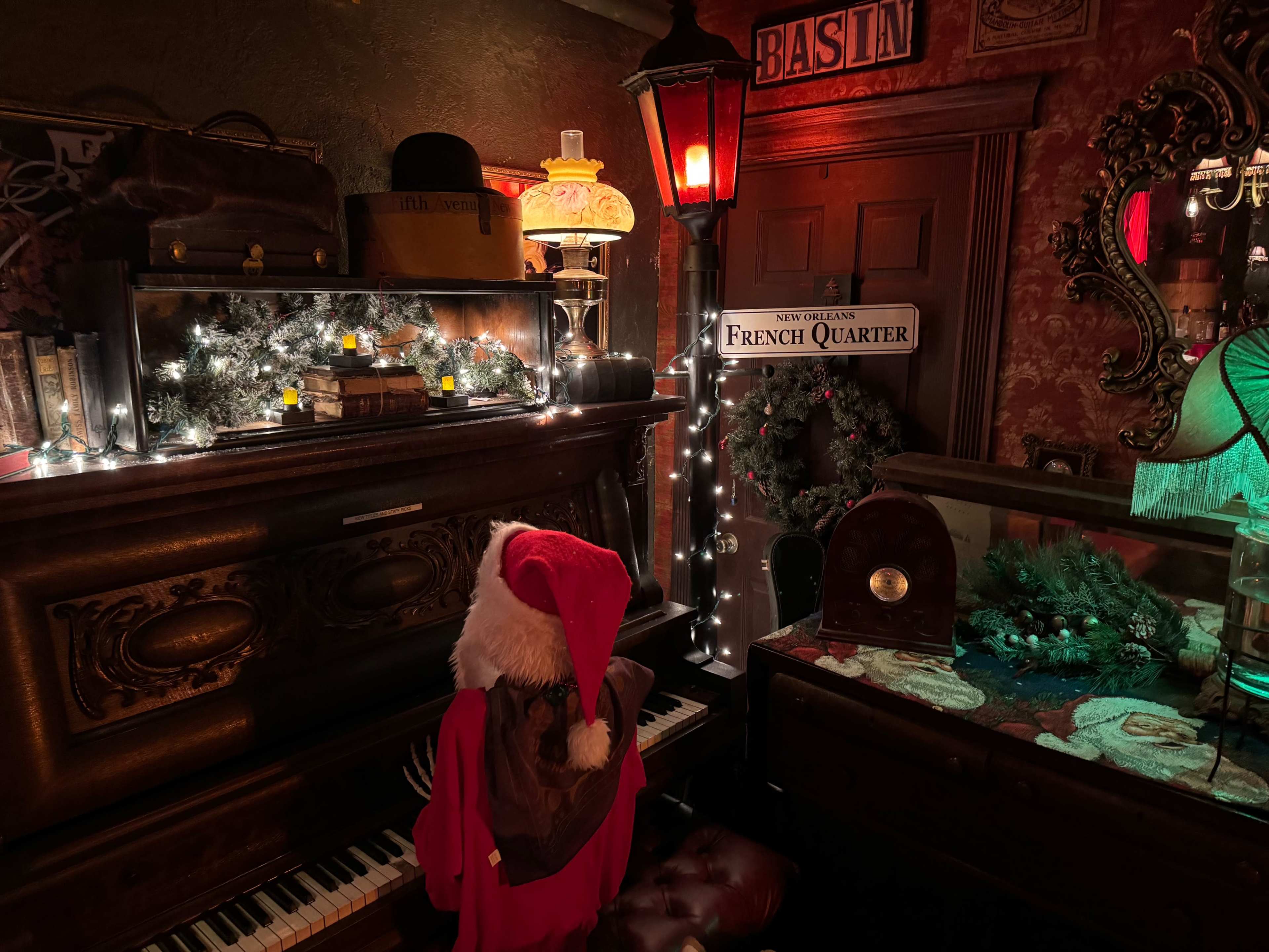 A person in a Santa hat plays a vintage piano surrounded by holiday decorations and dim lighting in a cozy room.