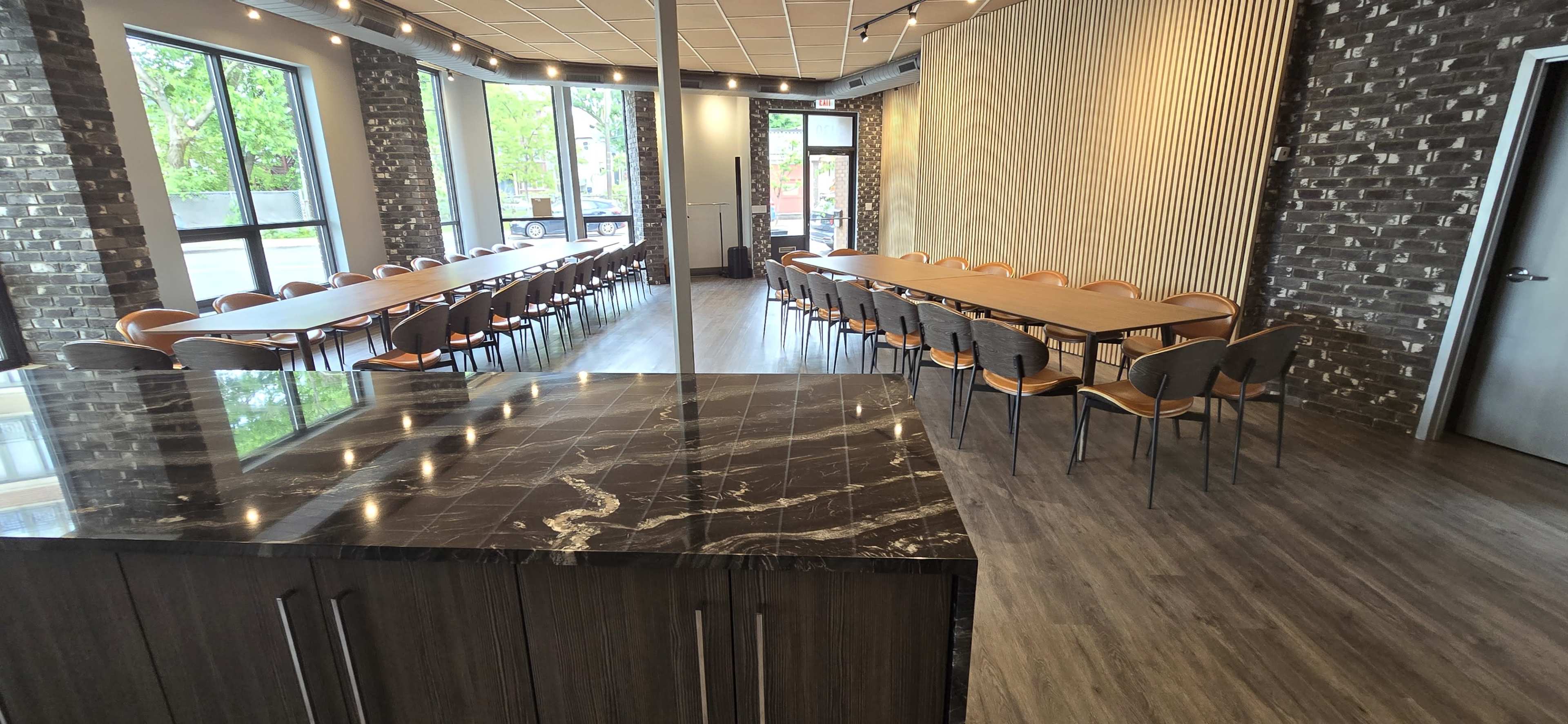 A spacious event room features long tables arranged in two parallel rows, with large windows allowing natural light to enter.