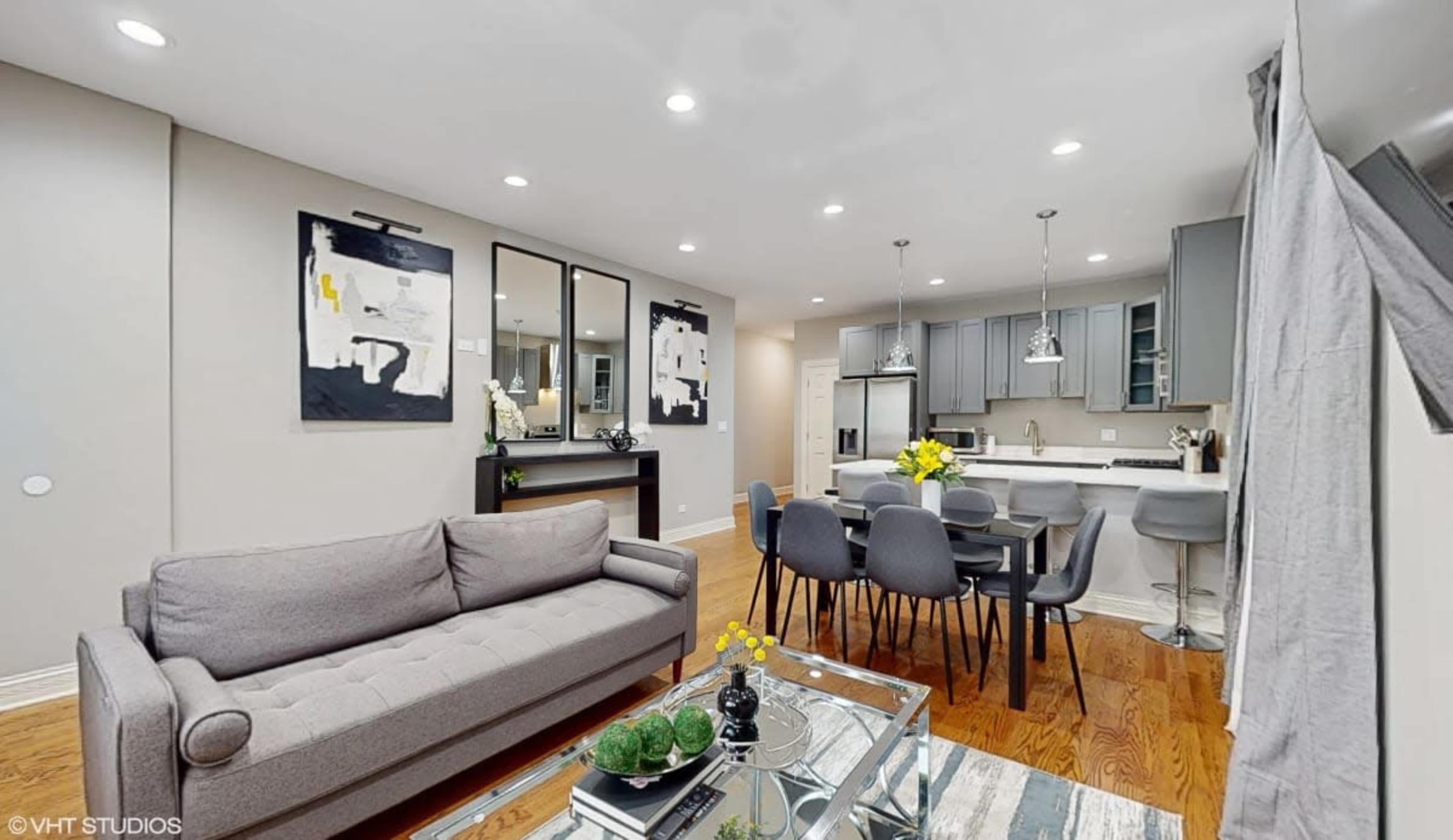 The image shows a modern living and dining area featuring a gray sofa, a glass coffee table with decor, and a dining table with gray chairs, all set in a well-lit, open space with contemporary artwork on the walls.