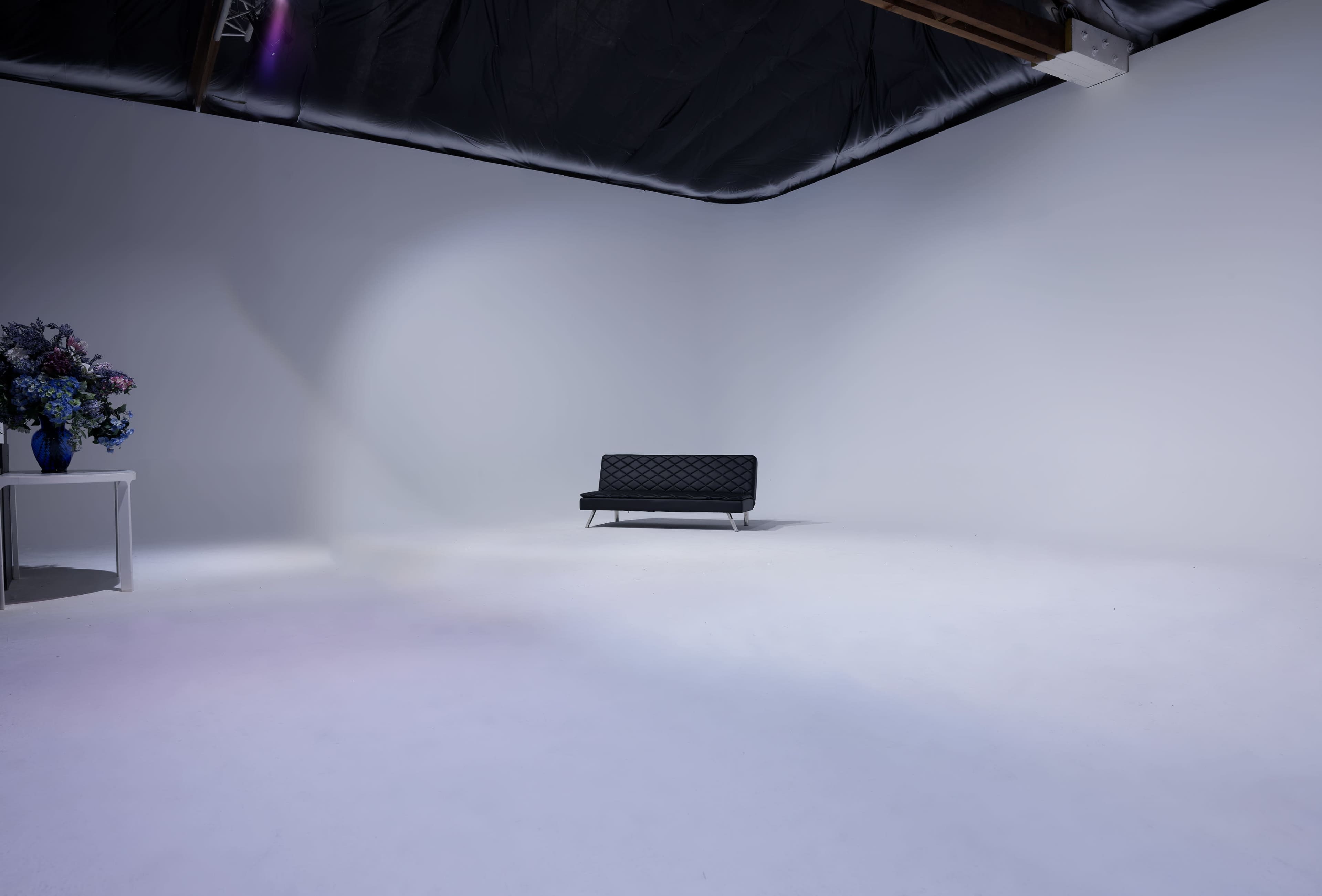 A black couch is positioned in the center of a sparsely furnished white room with a flower arrangement on a small table.
