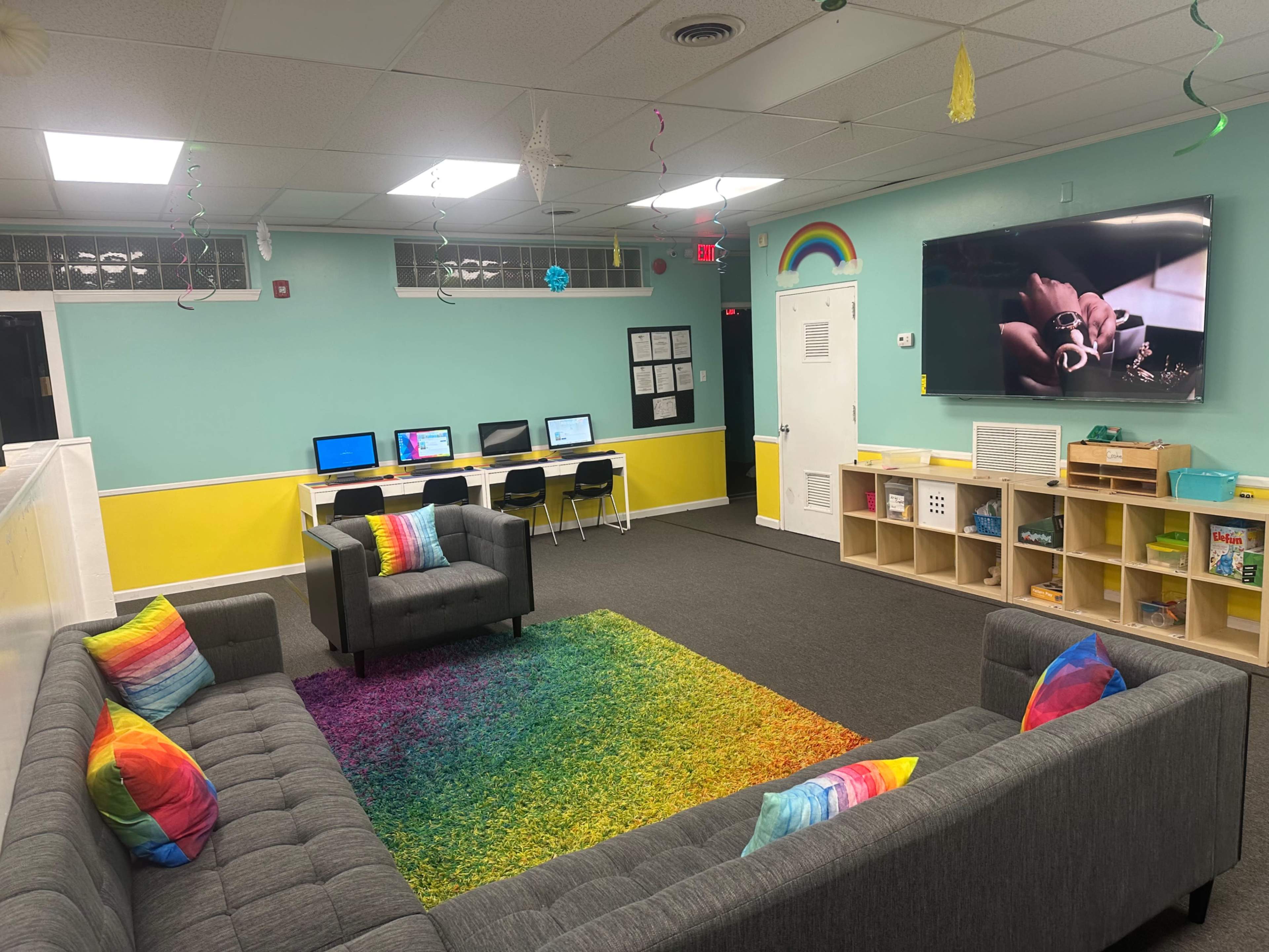 A brightly colored lounge area with a gray couch and chairs, a rainbow rug, computers along the wall, and a large screen displaying a video.