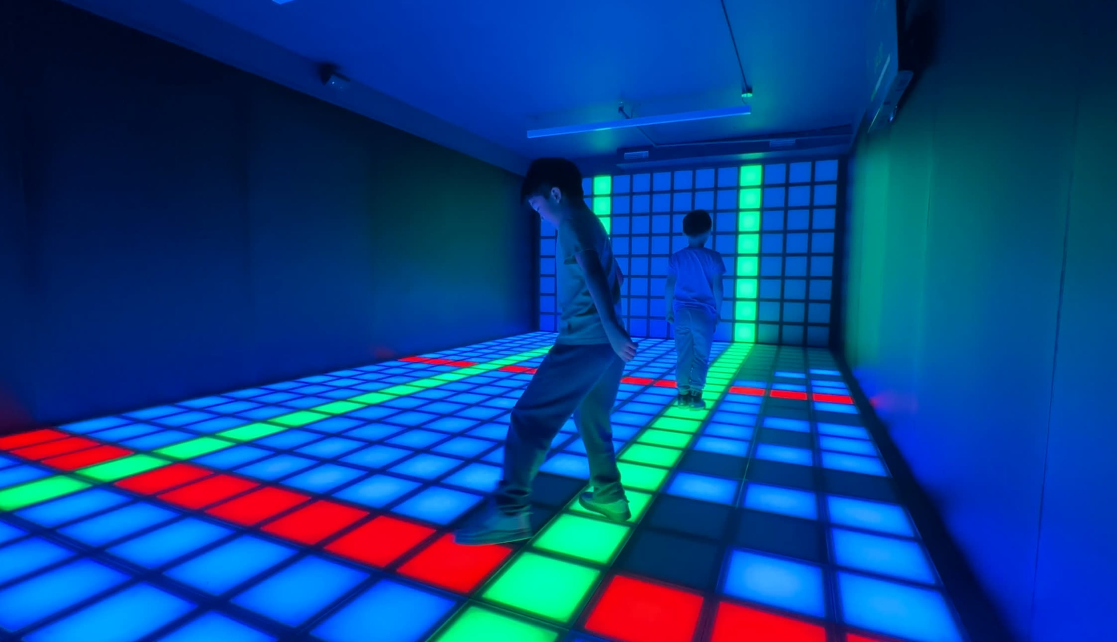 Two individuals walk on a brightly lit grid floor with blue, green, and red squares in a dimly lit room.