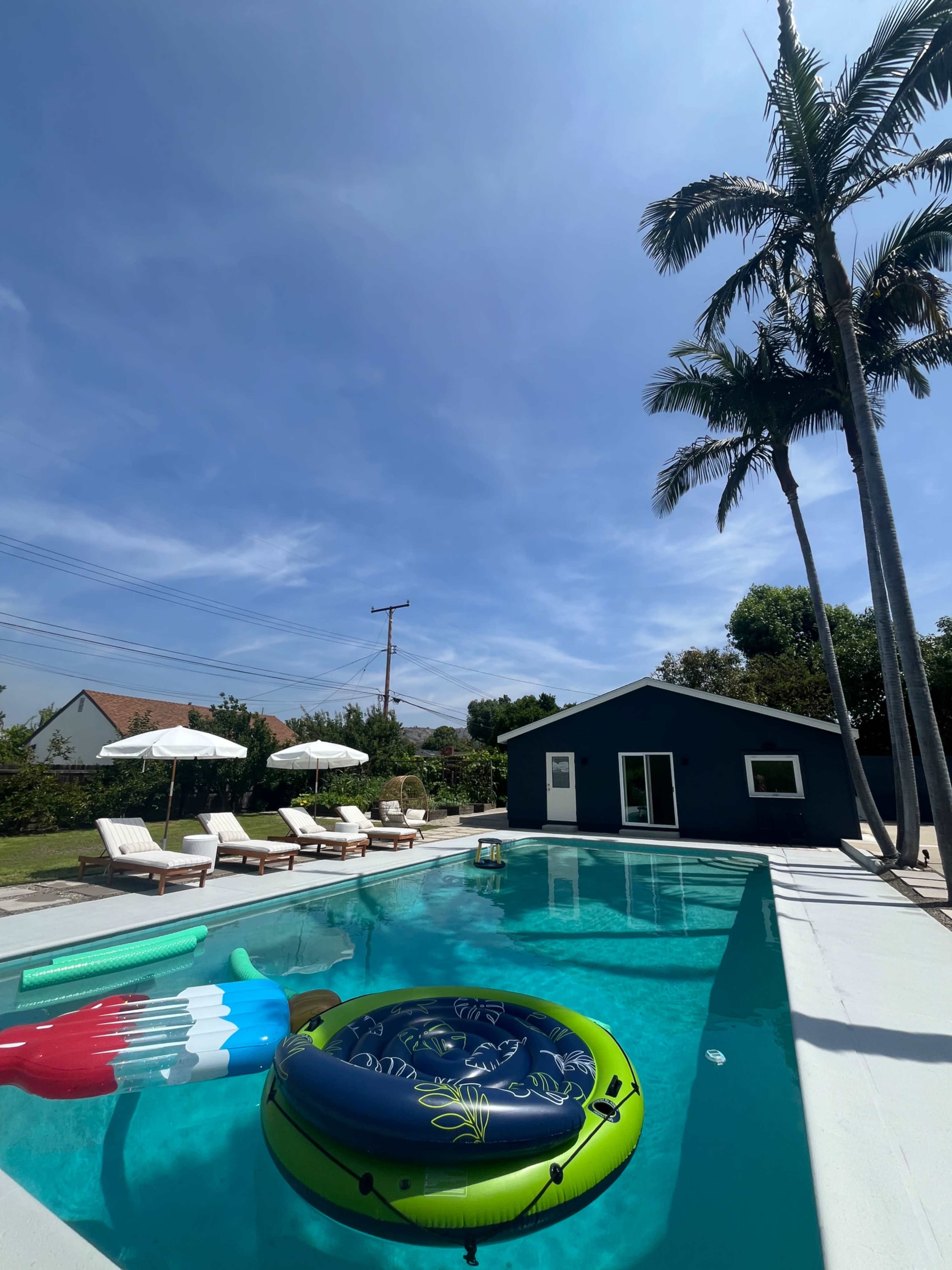 A swimming pool with a colorful float and lounge chairs is surrounded by palm trees and a nearby blue shed under a clear sky.