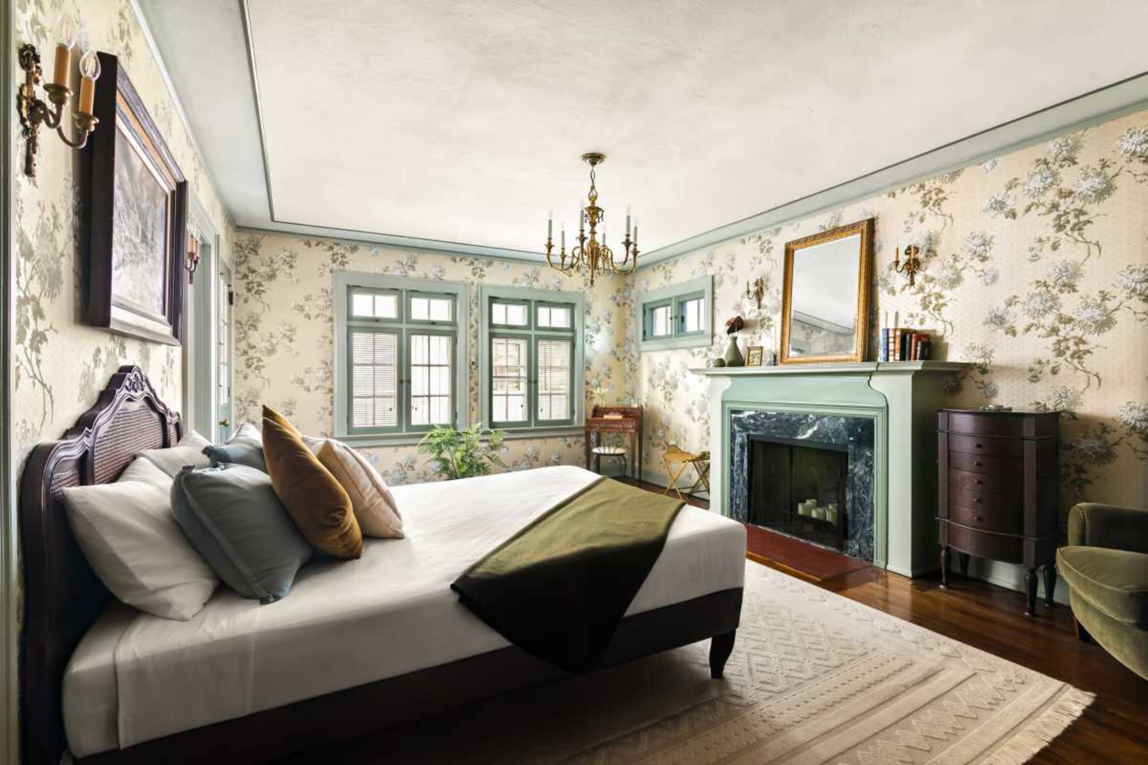 The image shows a neatly arranged bedroom featuring a bed, a fireplace, and several windows adorned with floral wallpaper.
