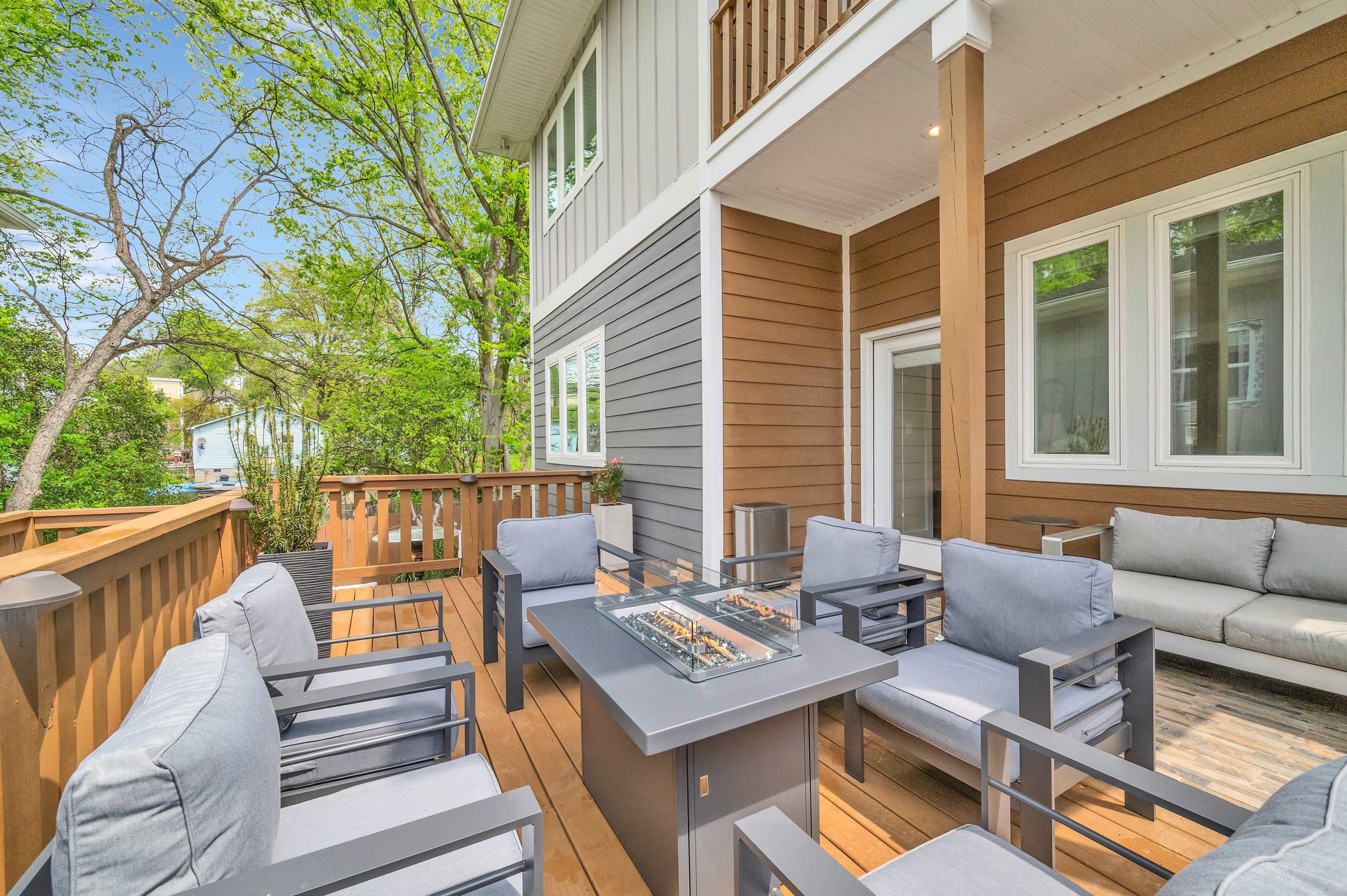 A spacious deck features modern seating and a firepit, surrounded by greenery and neighboring buildings.
