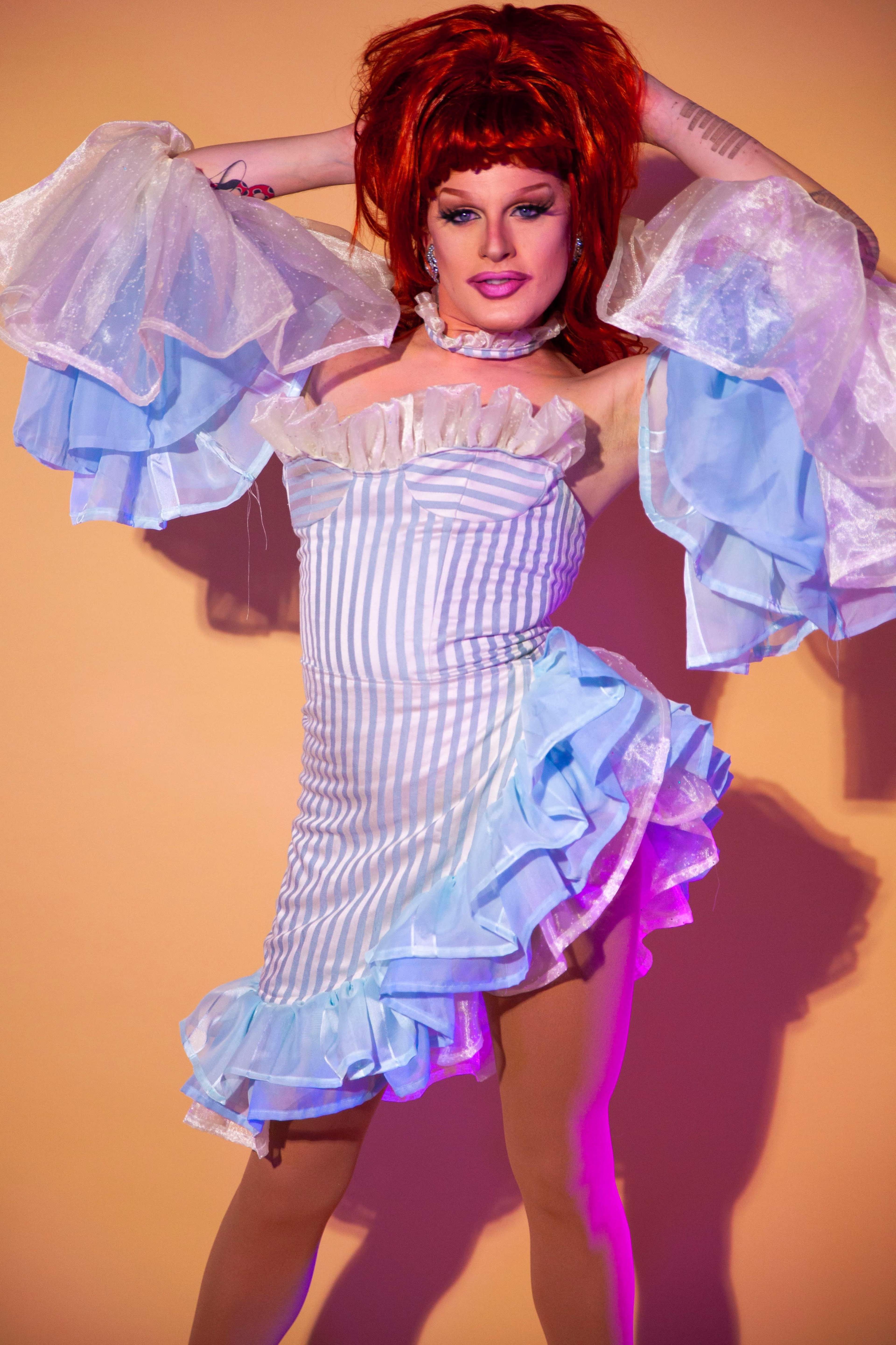 A performer poses in a striped dress with ruffled details against a soft orange background.