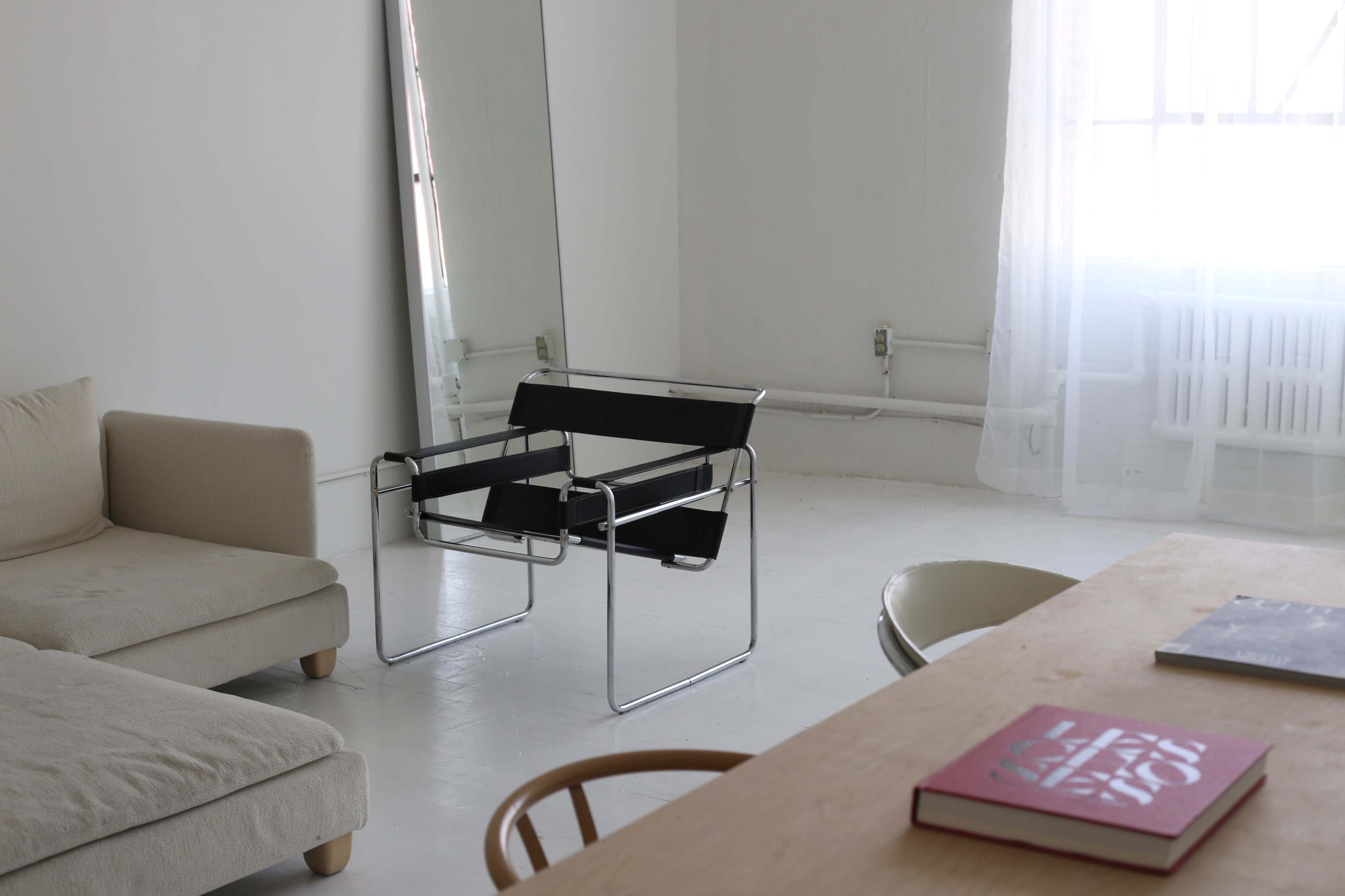 The image shows a minimalist room with a black chair, a beige couch, a wooden table with a book on it, and a large mirror reflecting sunlight.