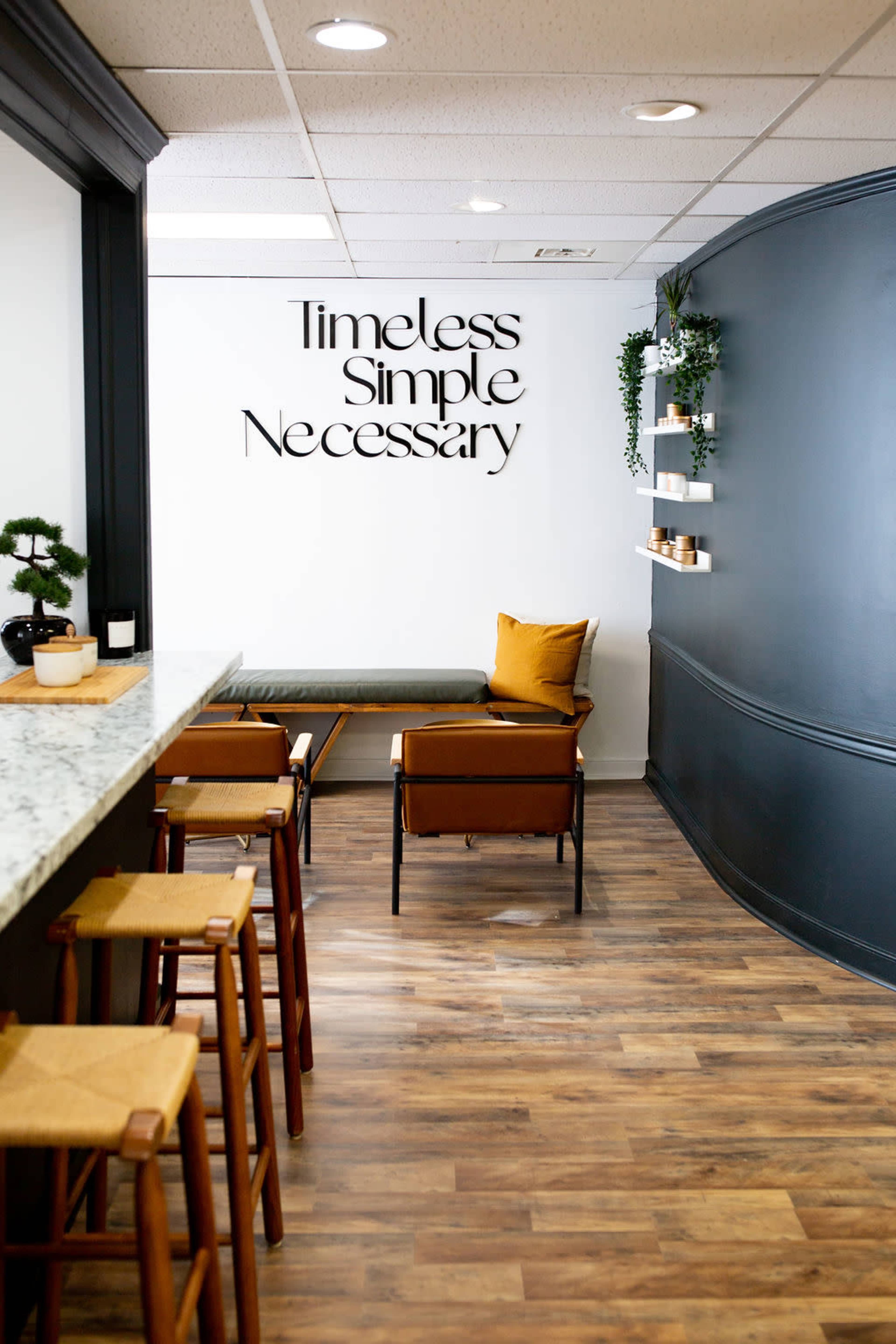 A minimalist interior design featuring a bar counter with stools, a bench, and a wall with the words "Timeless Simple Necessary" written in an artistic font.