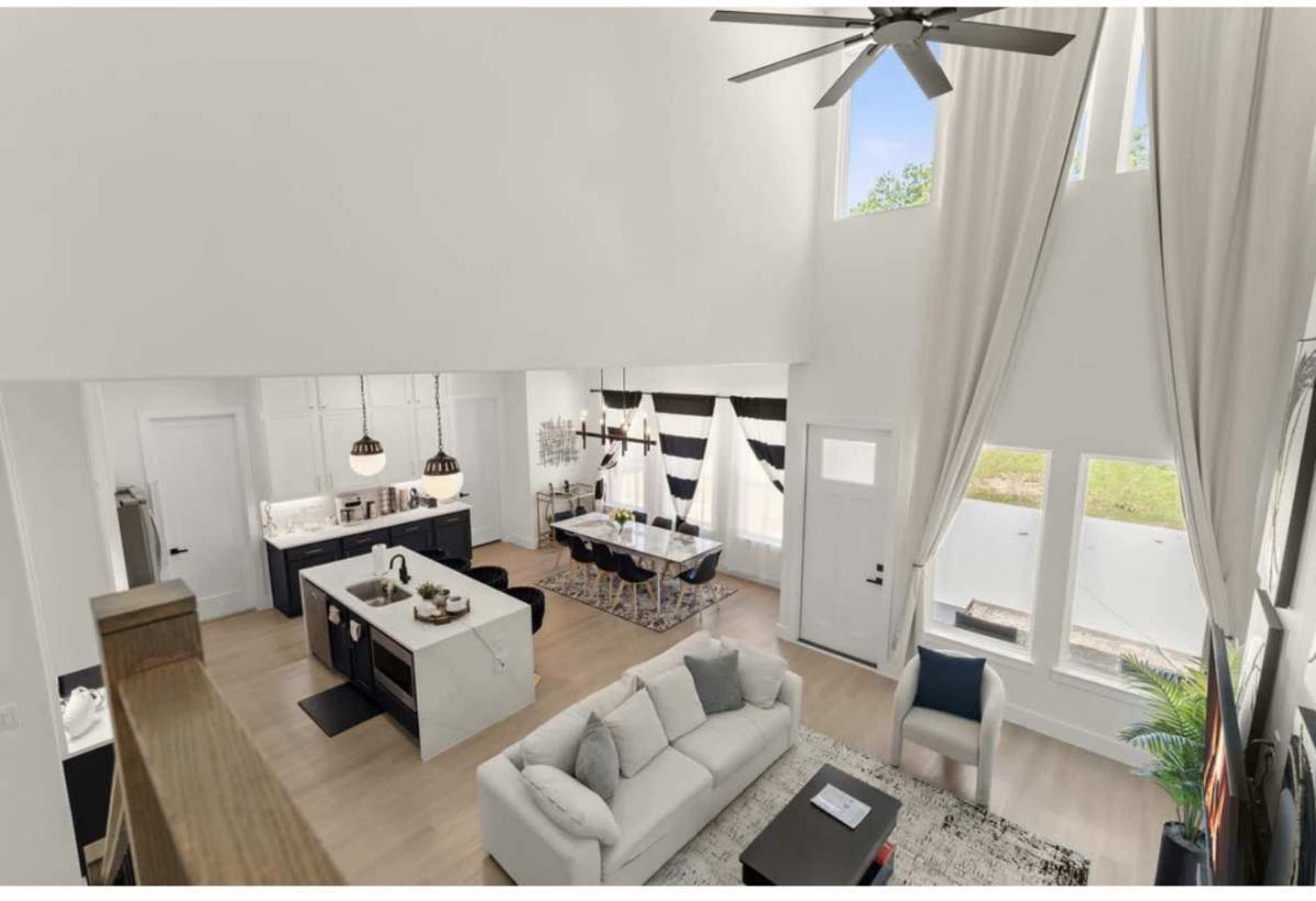 The image shows a modern living space featuring an open-concept design with a kitchen, dining area, and living room, illuminated by large windows and a high ceiling.