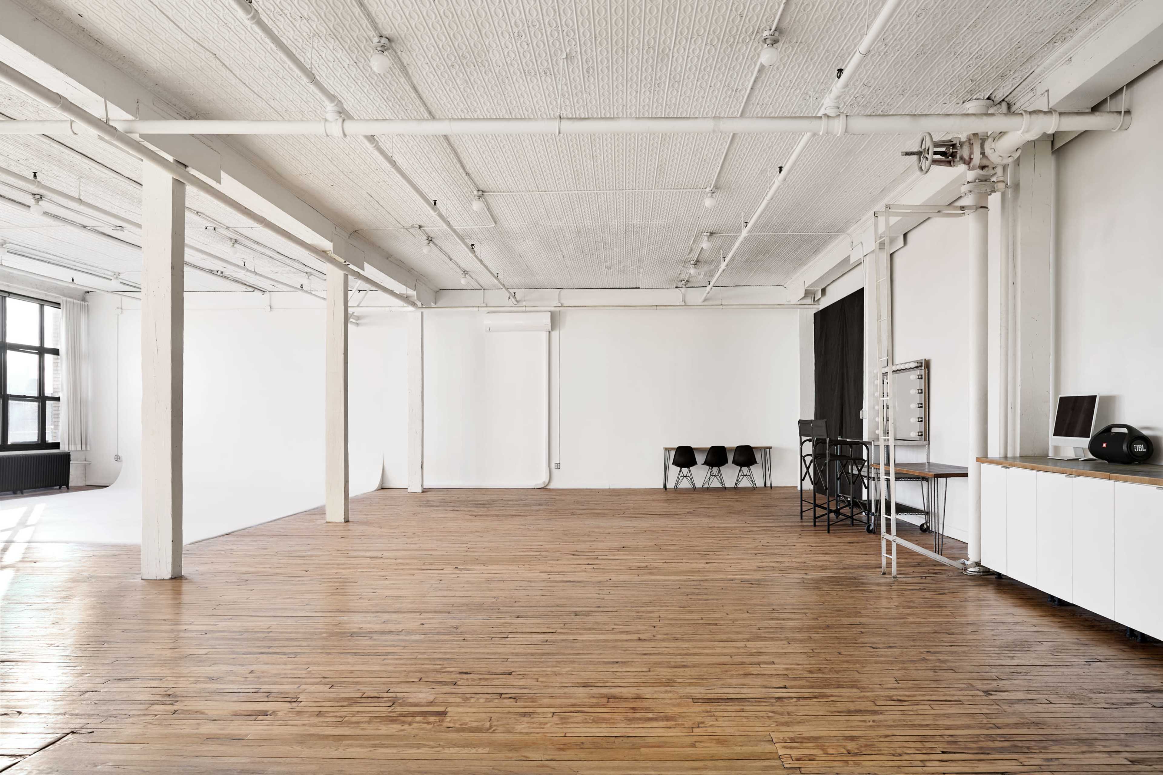 The image shows a spacious, empty industrial-style room with wooden floors, white walls, and minimal furnishings, including some chairs and a desk.