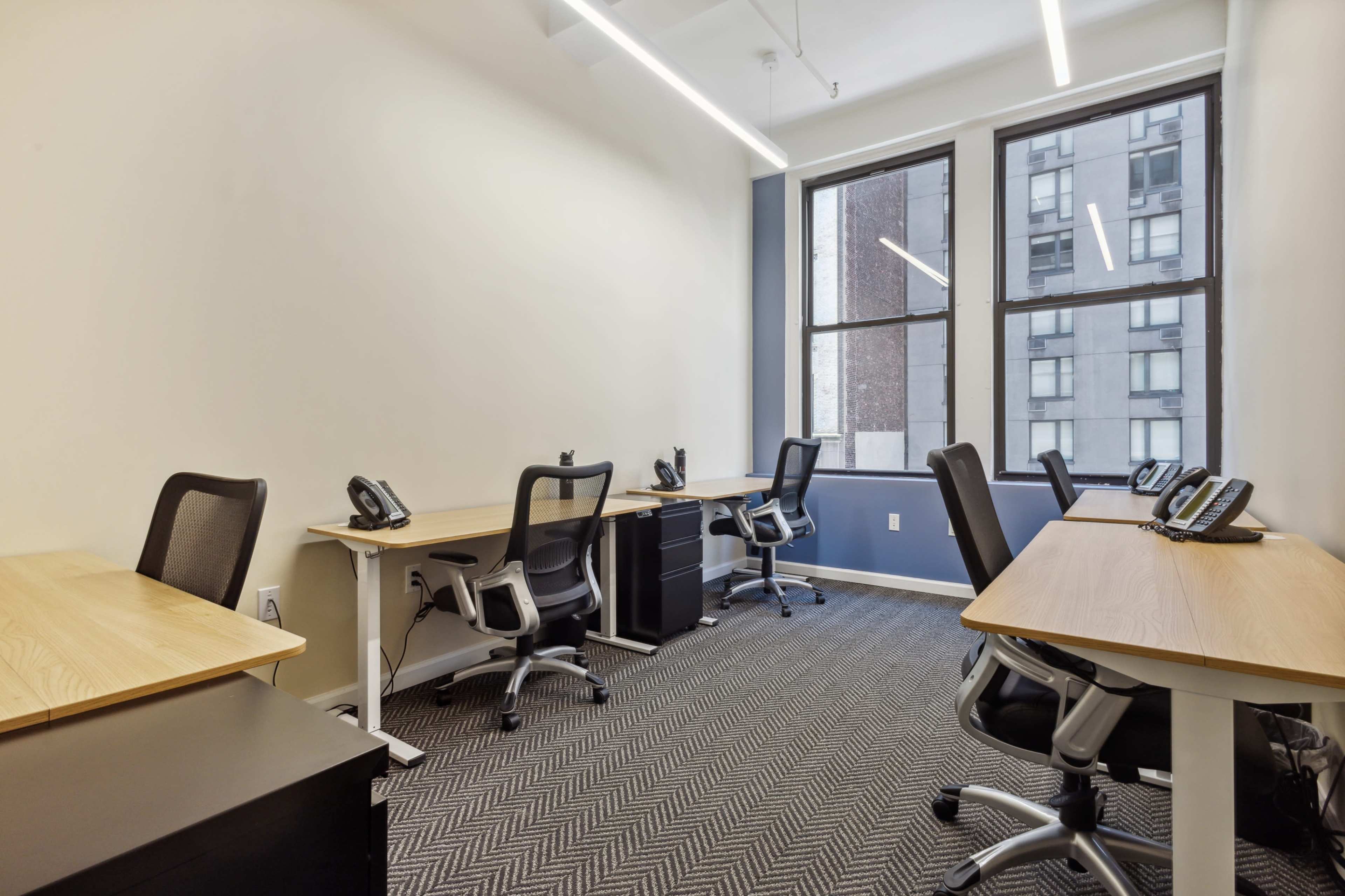 A modern office space with several desks and chairs, each equipped with a telephone, and large windows providing natural light.