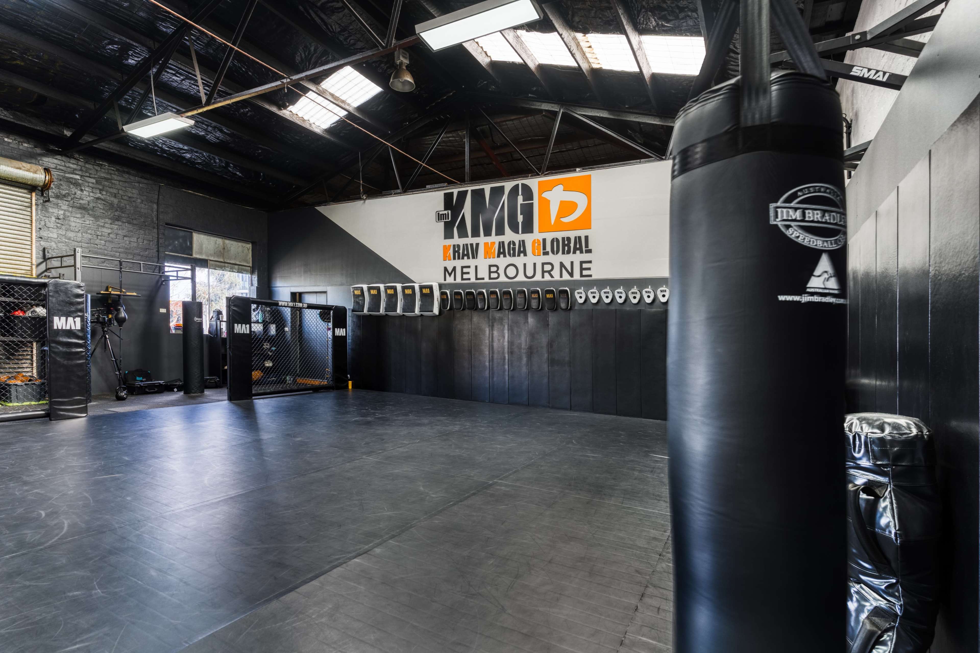 The image shows an empty martial arts training facility with boxing bags and a large wall mural displaying "KMG Krav Maga Global Melbourne."