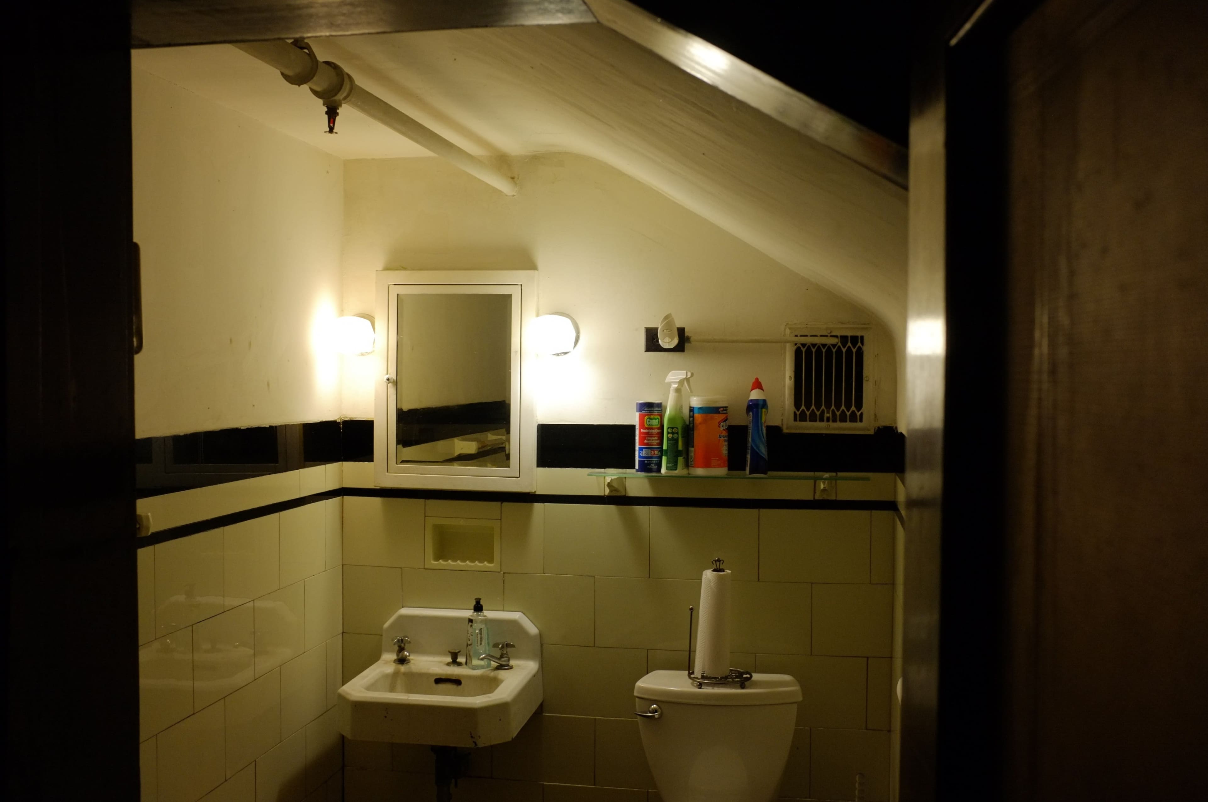 The image shows a small, sloped bathroom with a sink, toilet, and cleaning supplies on a shelf.