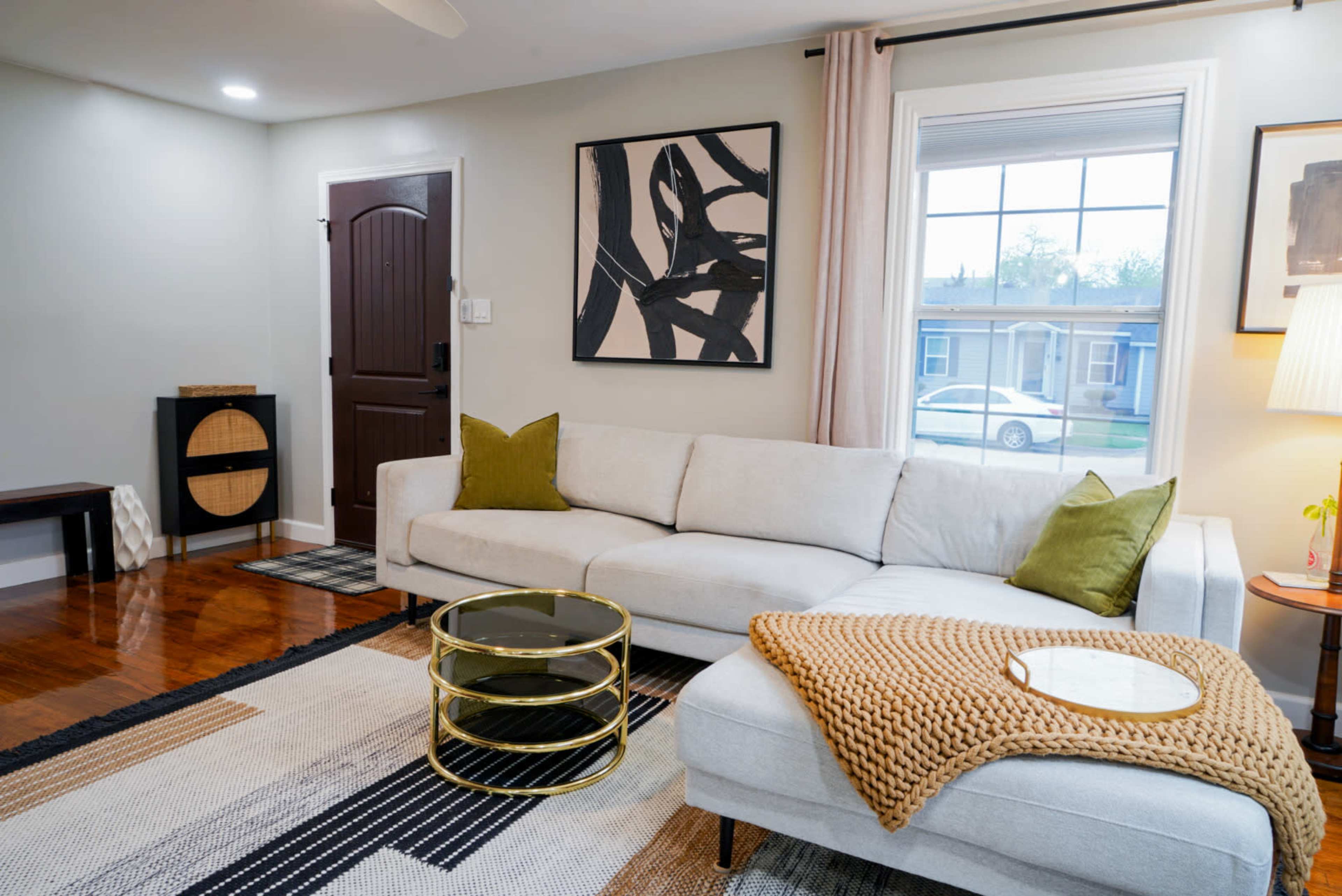 A modern living room features a light-colored sofa with green accent pillows, a coffee table, and a patterned rug, illuminated by natural light from a window.