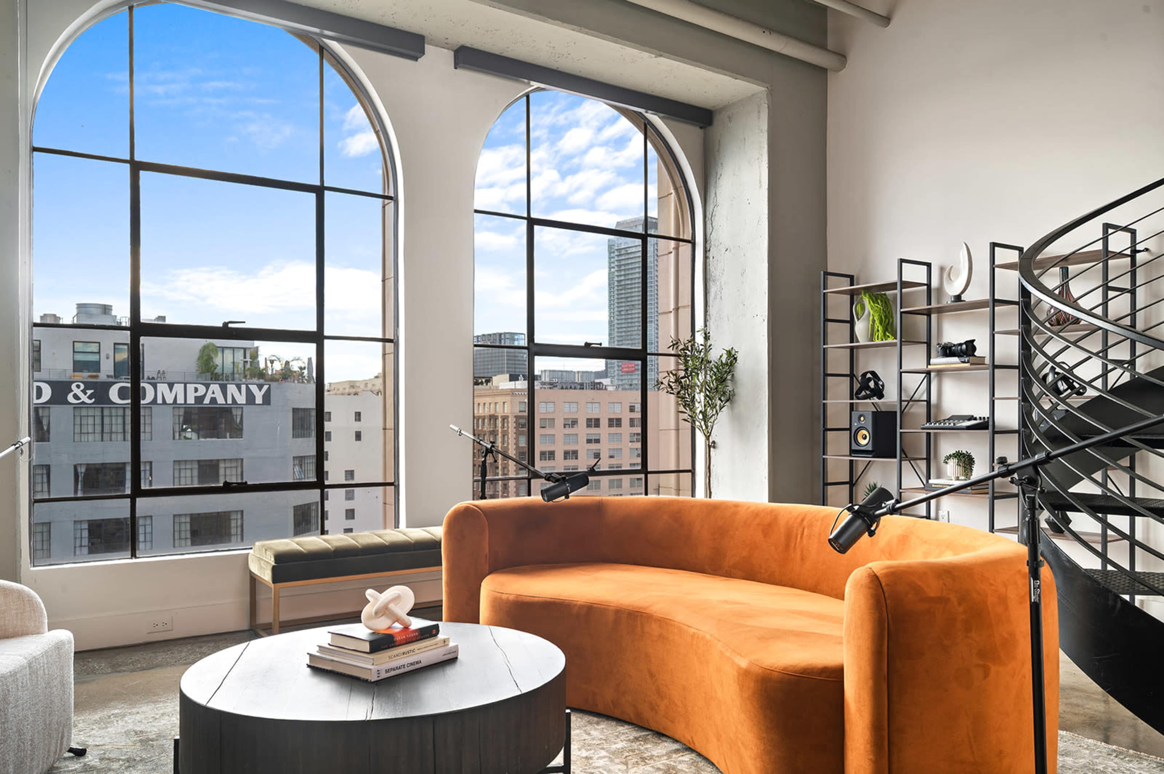 The scene features a modern interior with large arched windows allowing natural light to fill the space, showcasing a curved orange sofa, a round black table, and a spiral staircase in a loft-like setting.